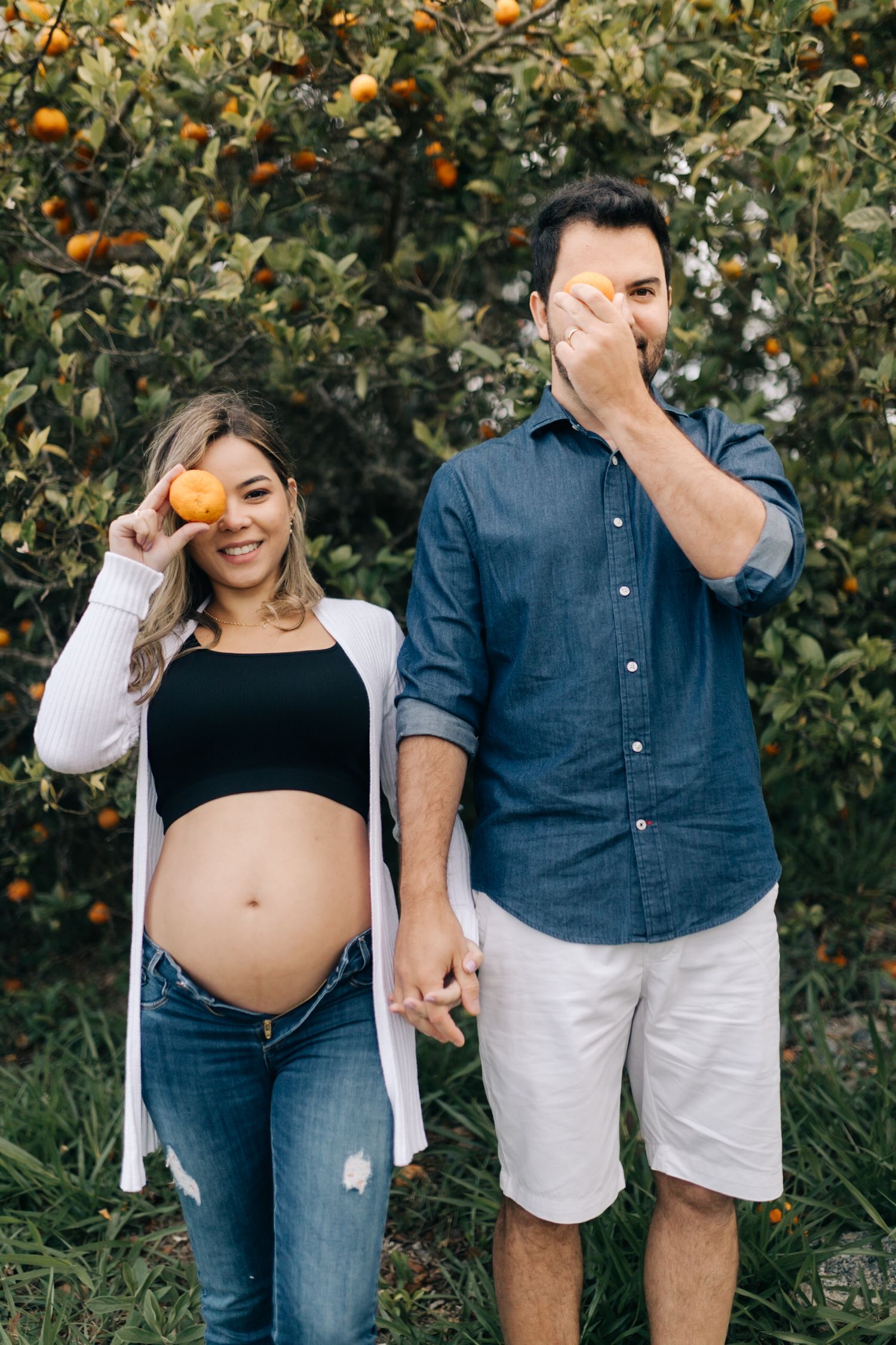 A couple holds oranges in front of their faces while standing in a citrus grove.