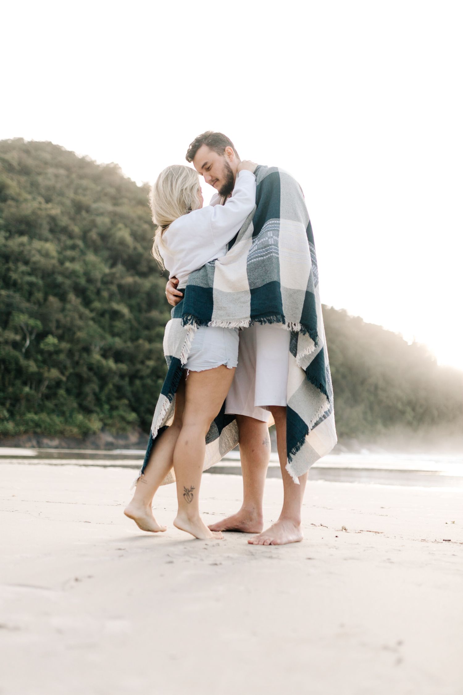 A couple shares an intimate moment on a misty beach while wrapped in a plaid blanket during sunset.