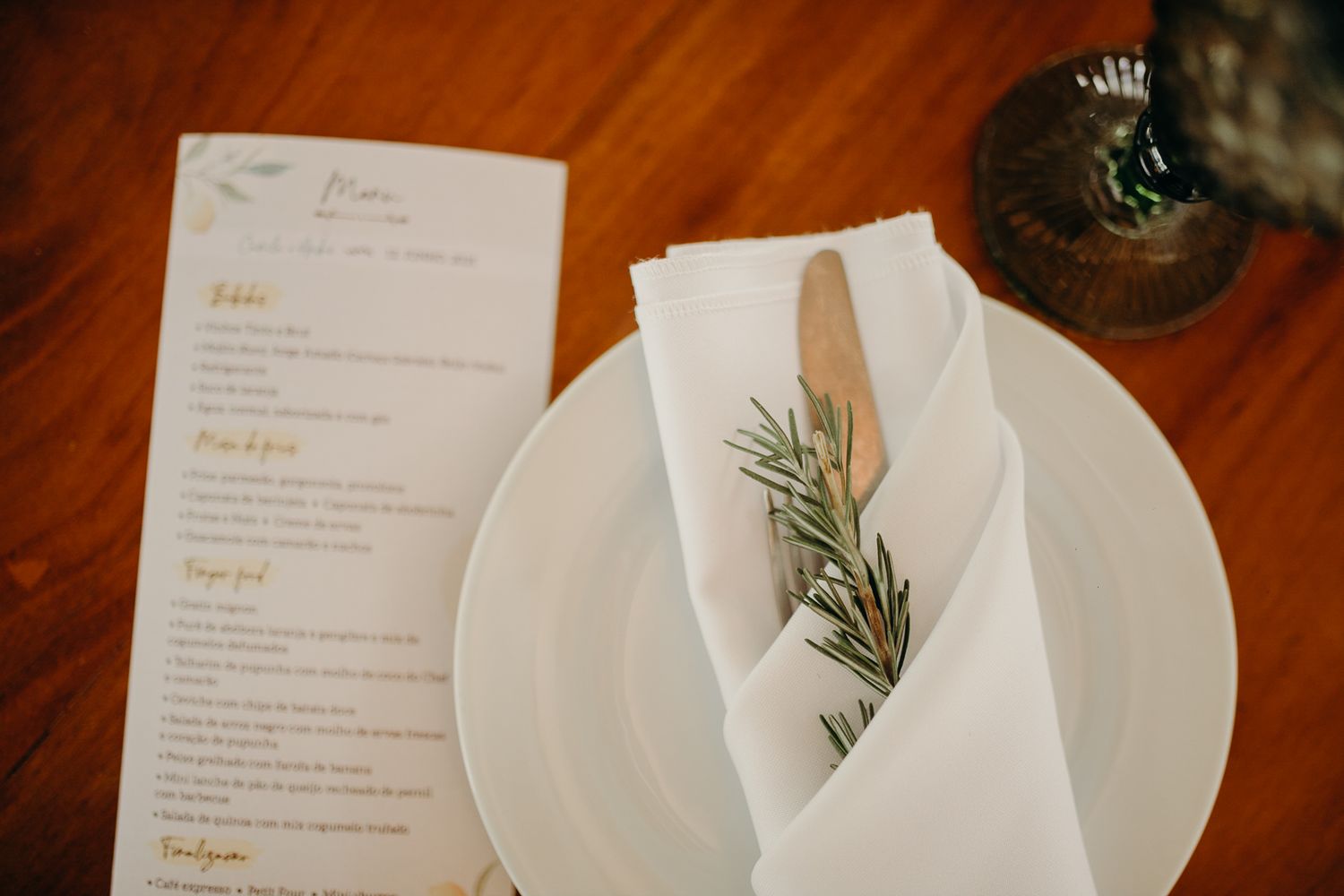 Elegant table setting with white napkin, rosemary sprig, and menu on wooden table.