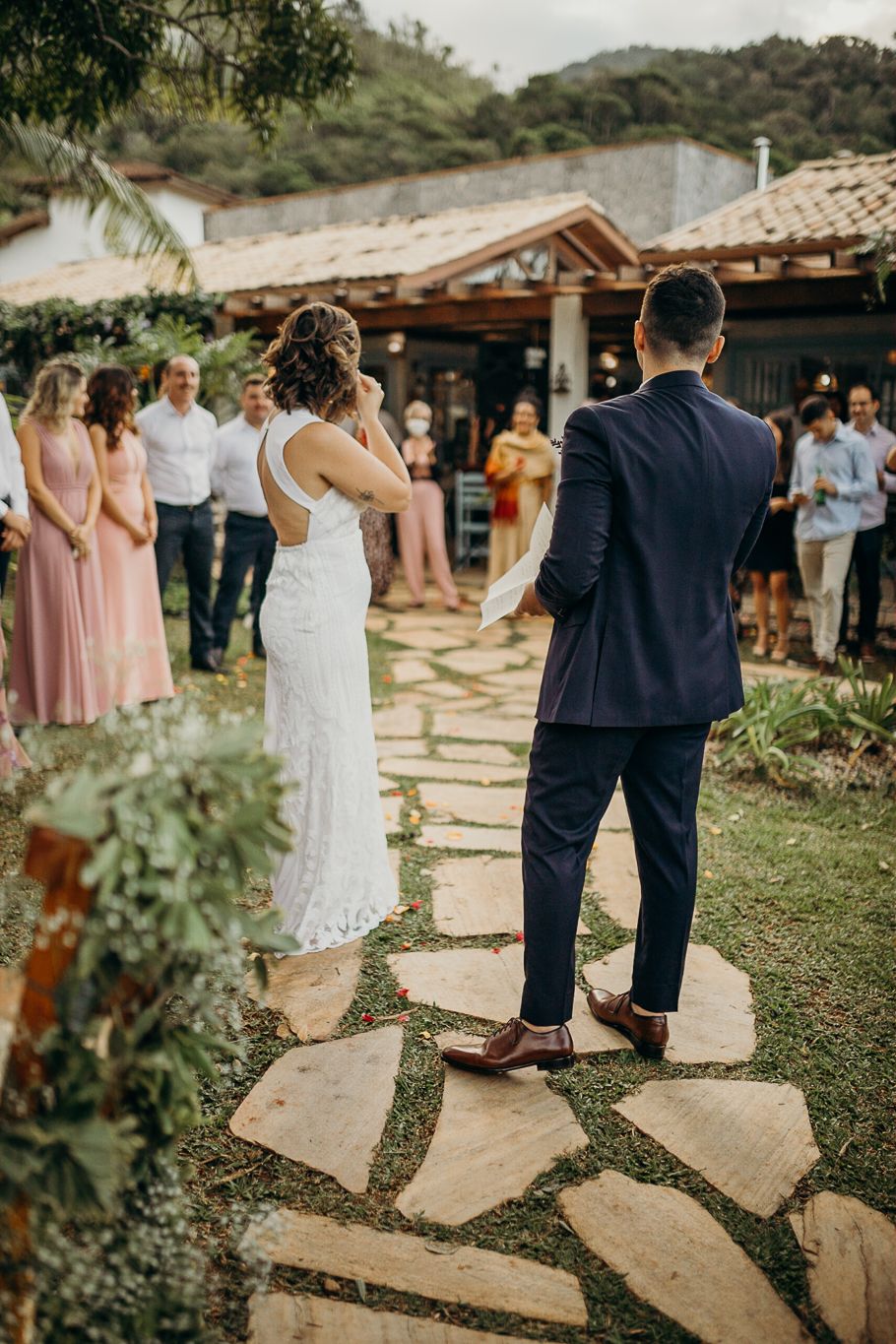 A couple stands on a stone path during their outdoor wedding ceremony with guests watching in the background.
