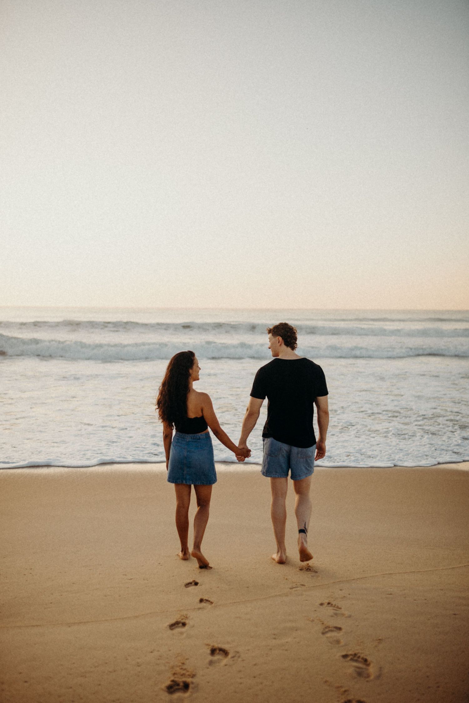 Couple leaves footprints in sand while walking along ocean shoreline at sunset.