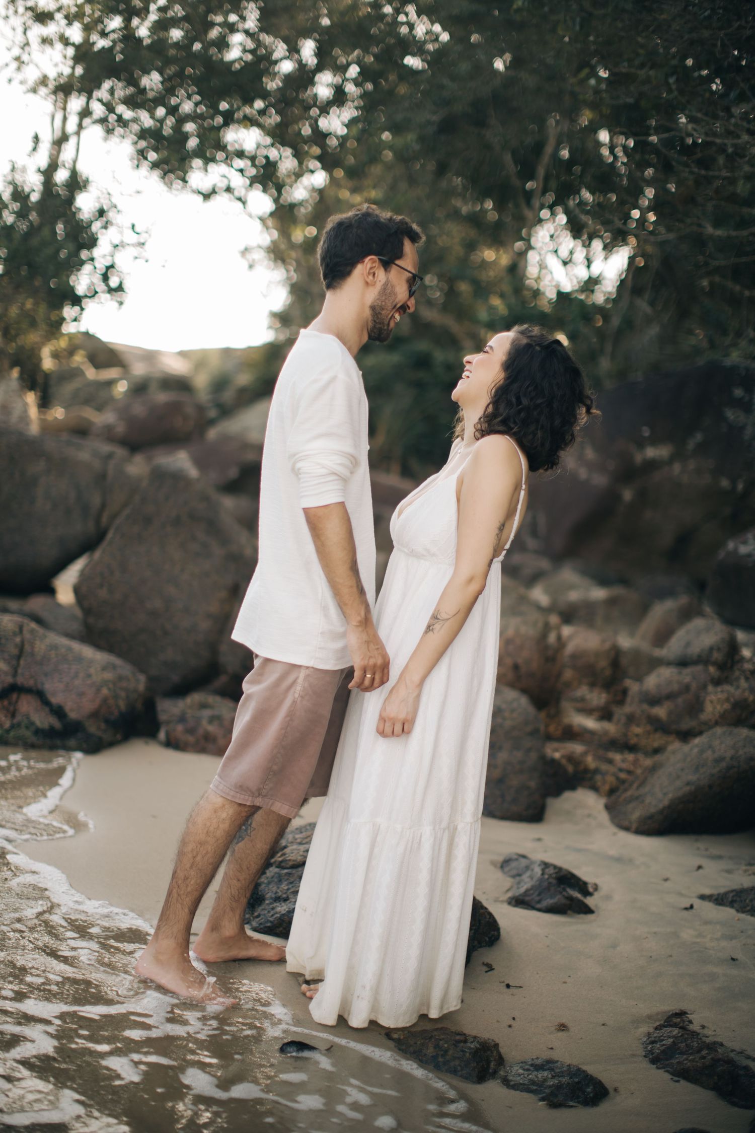 Couple standing in shallow water on rocky beach at sunset in matching white attire.