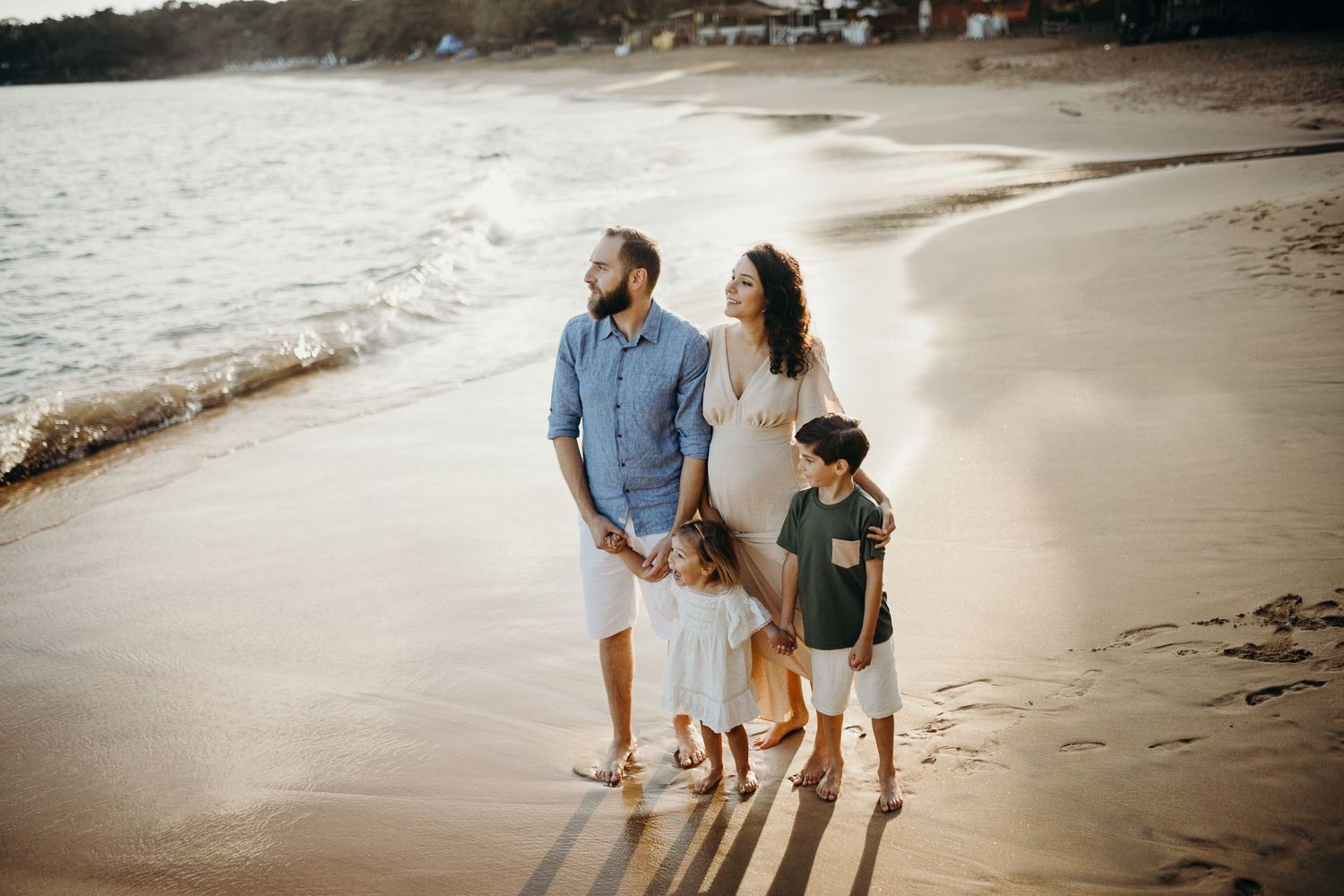 Family walking along shoreline during sunset with long shadows on sandy beach.