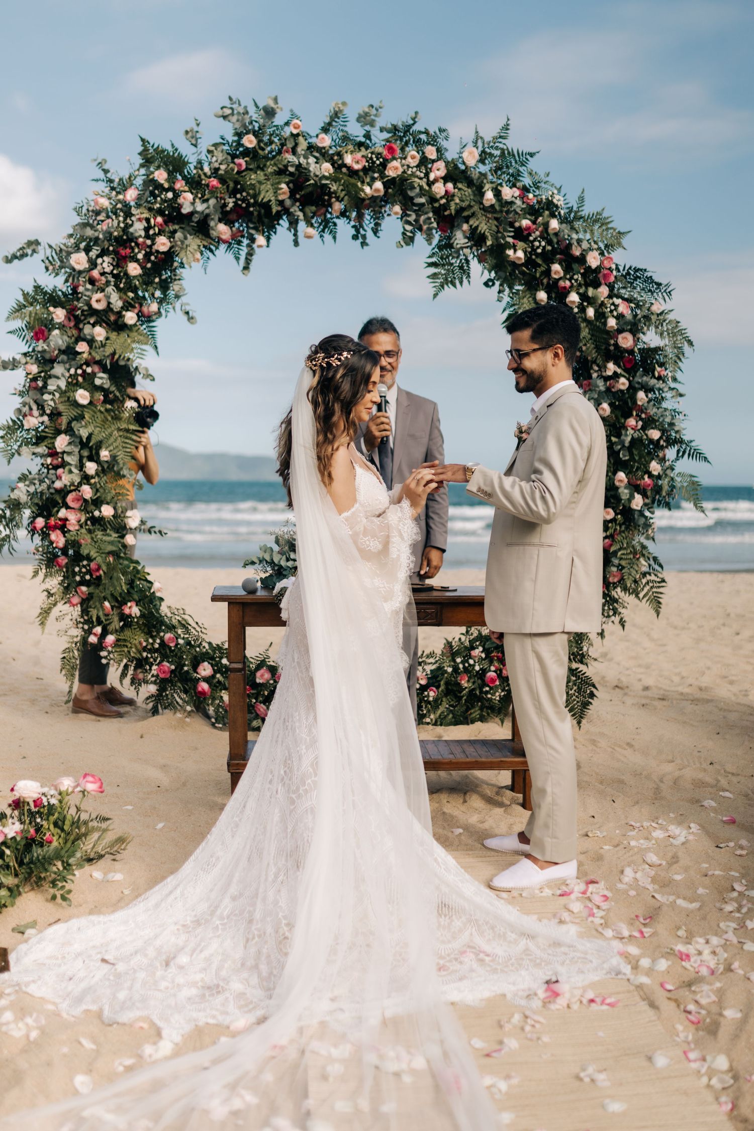 A couple exchanges rings during their beach wedding ceremony under a flower-adorned arch.