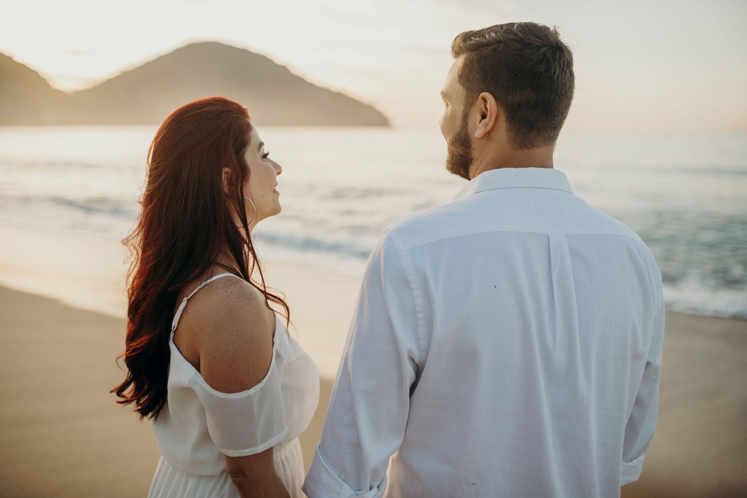 Romantic couple in white outfits standing on beach at sunset with mountains in background.