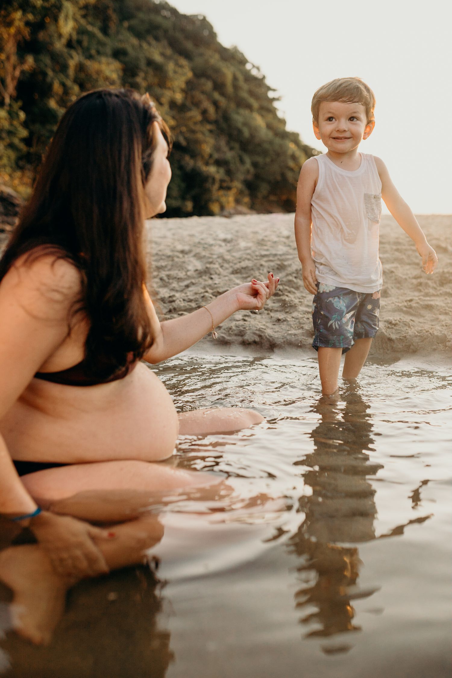 Woman in swimwear sits in shallow water while child stands and plays during a beach sunset.