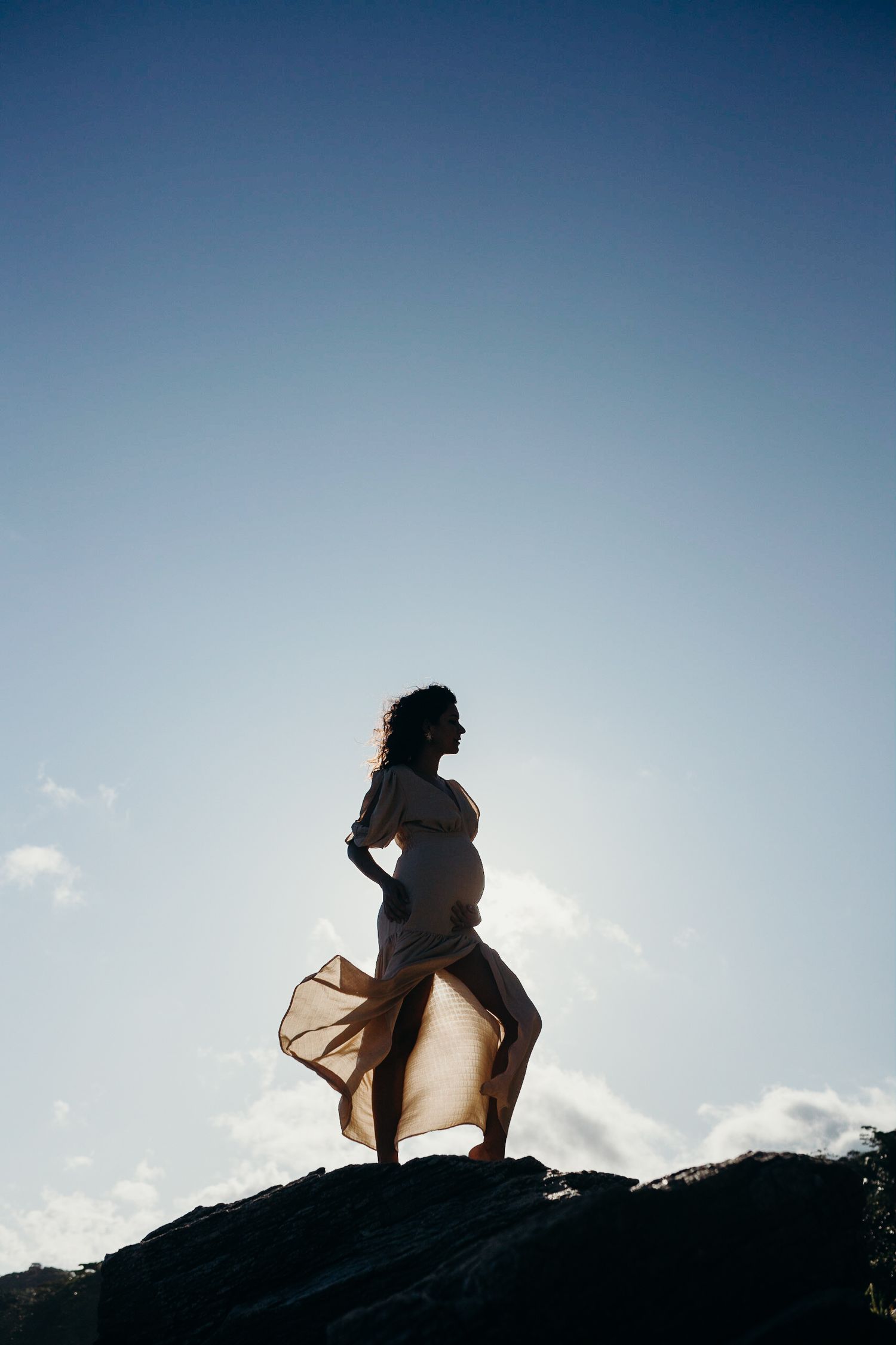 Side profile of pregnant silhouette on rocky cliff with dress flowing in breeze.
