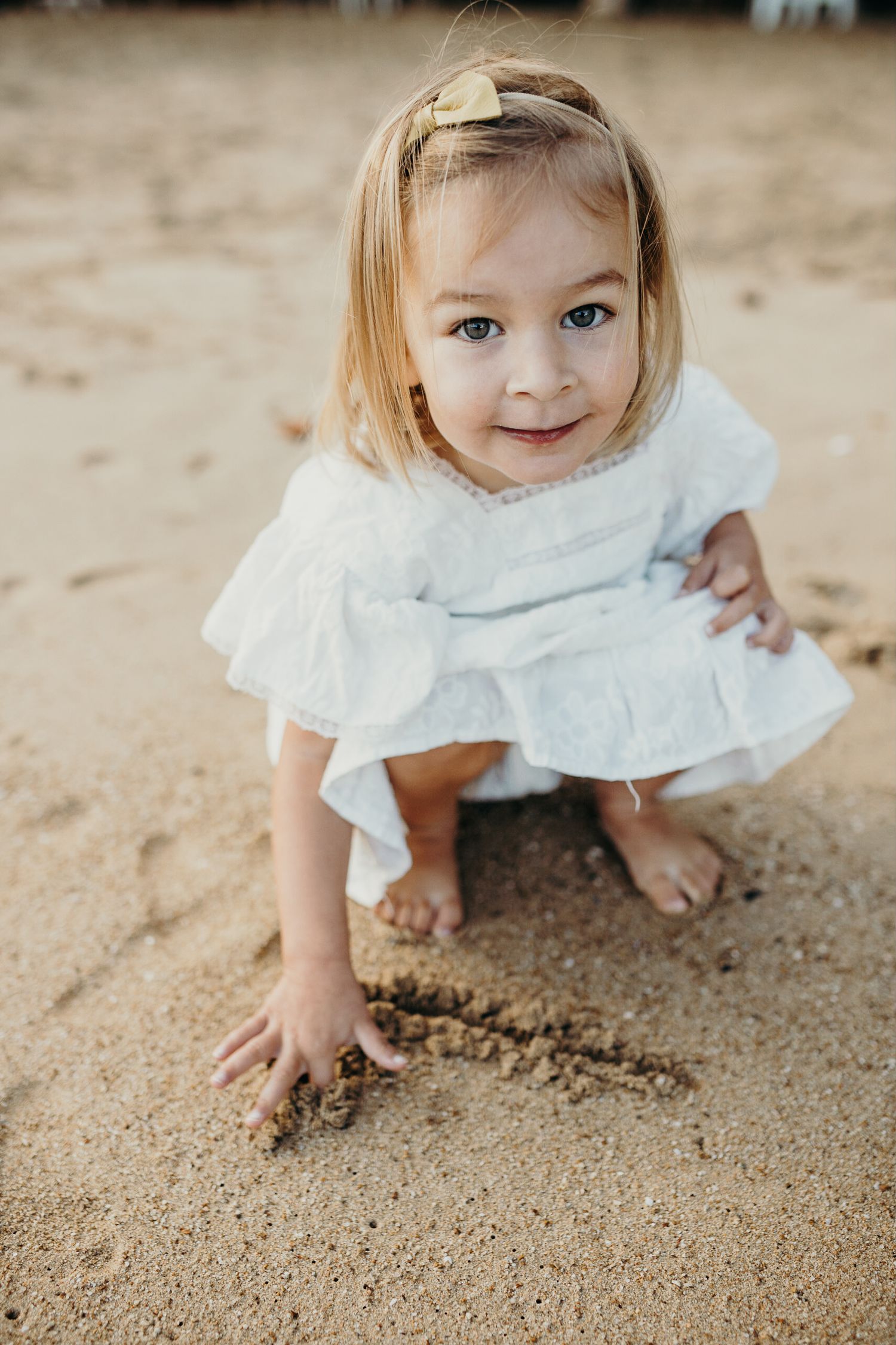 A small child in white dress plays in the sand at the beach.