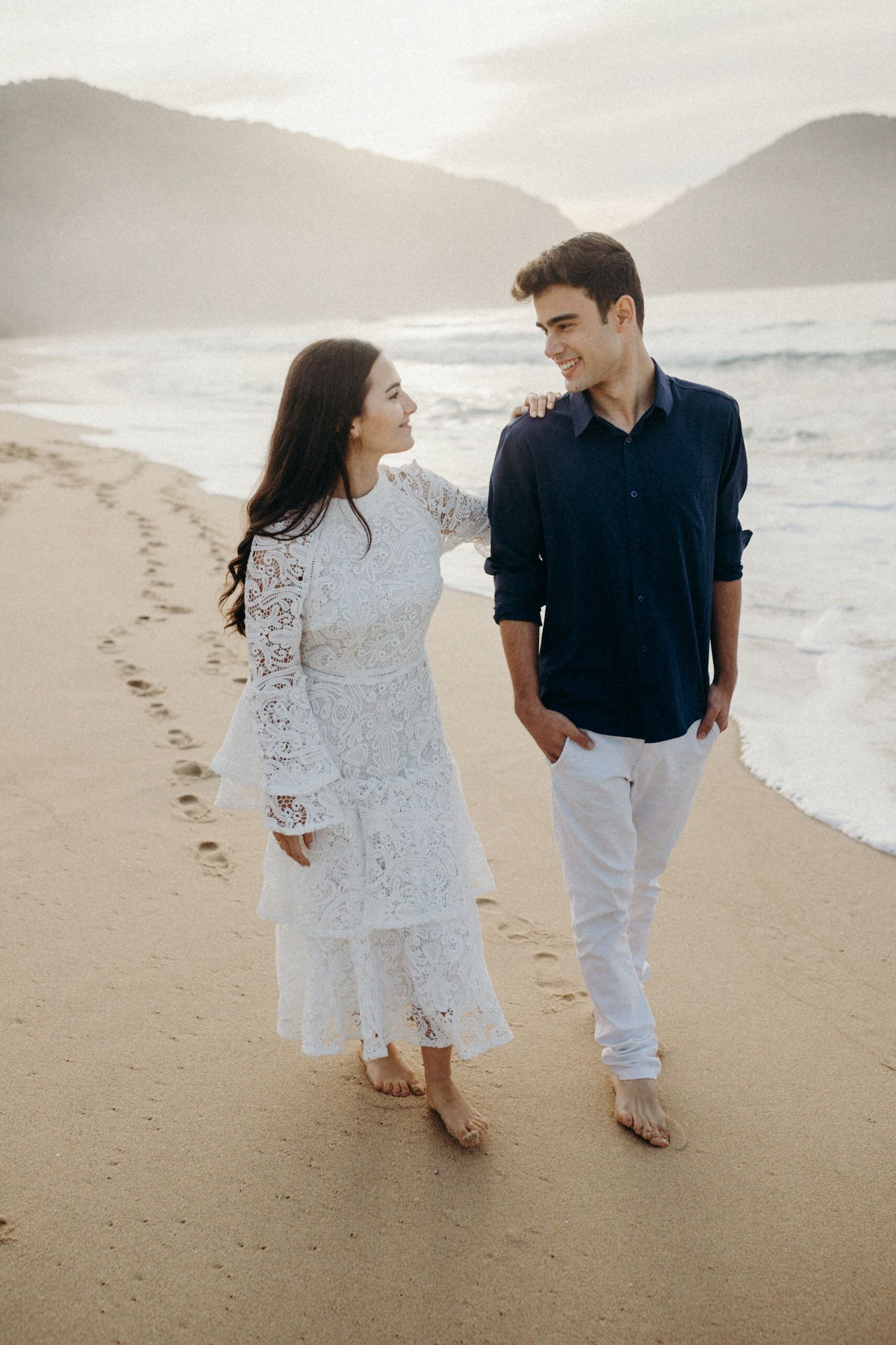 A couple walks together on a sandy beach wearing coordinated white and navy outfits against a misty backdrop.