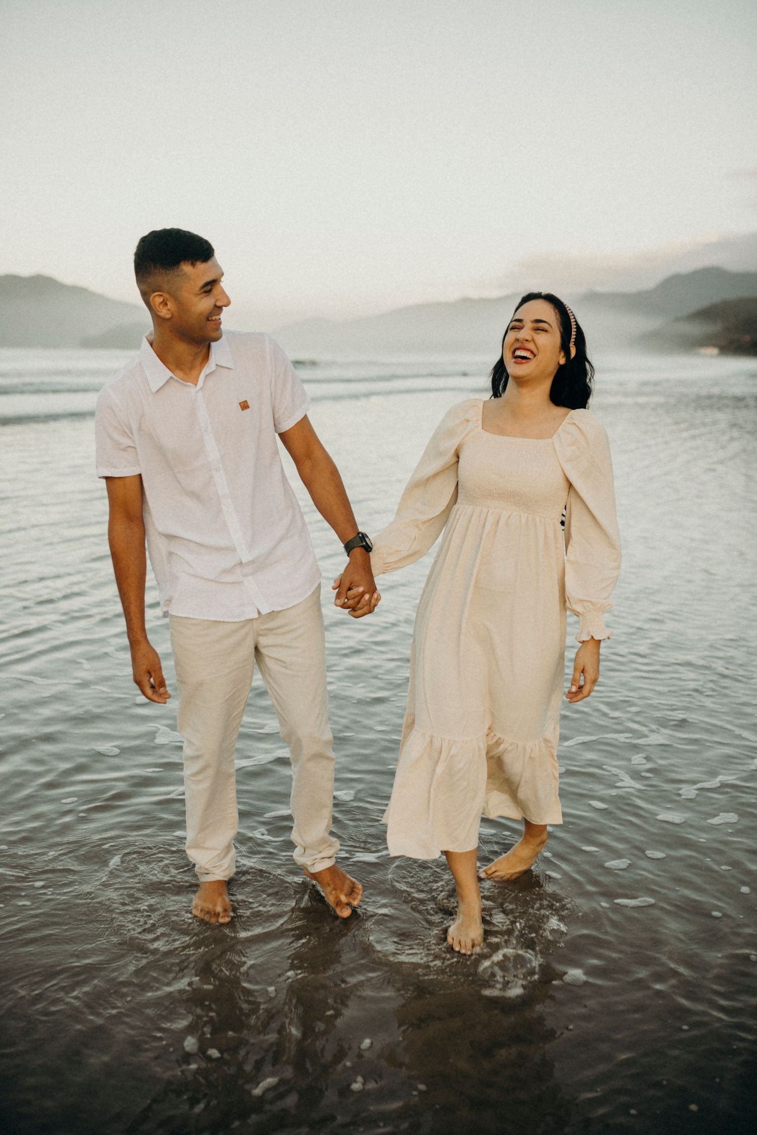Joyful couple laughing while walking hand in hand in shallow ocean water at sunset.