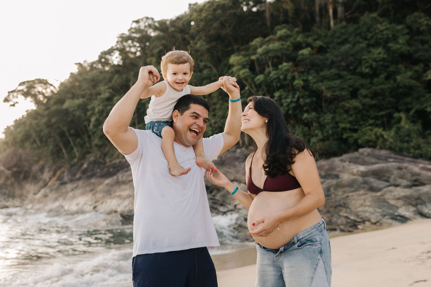 Joyful family moment on a scenic beach with rocky cliffs and lush greenery in the background.