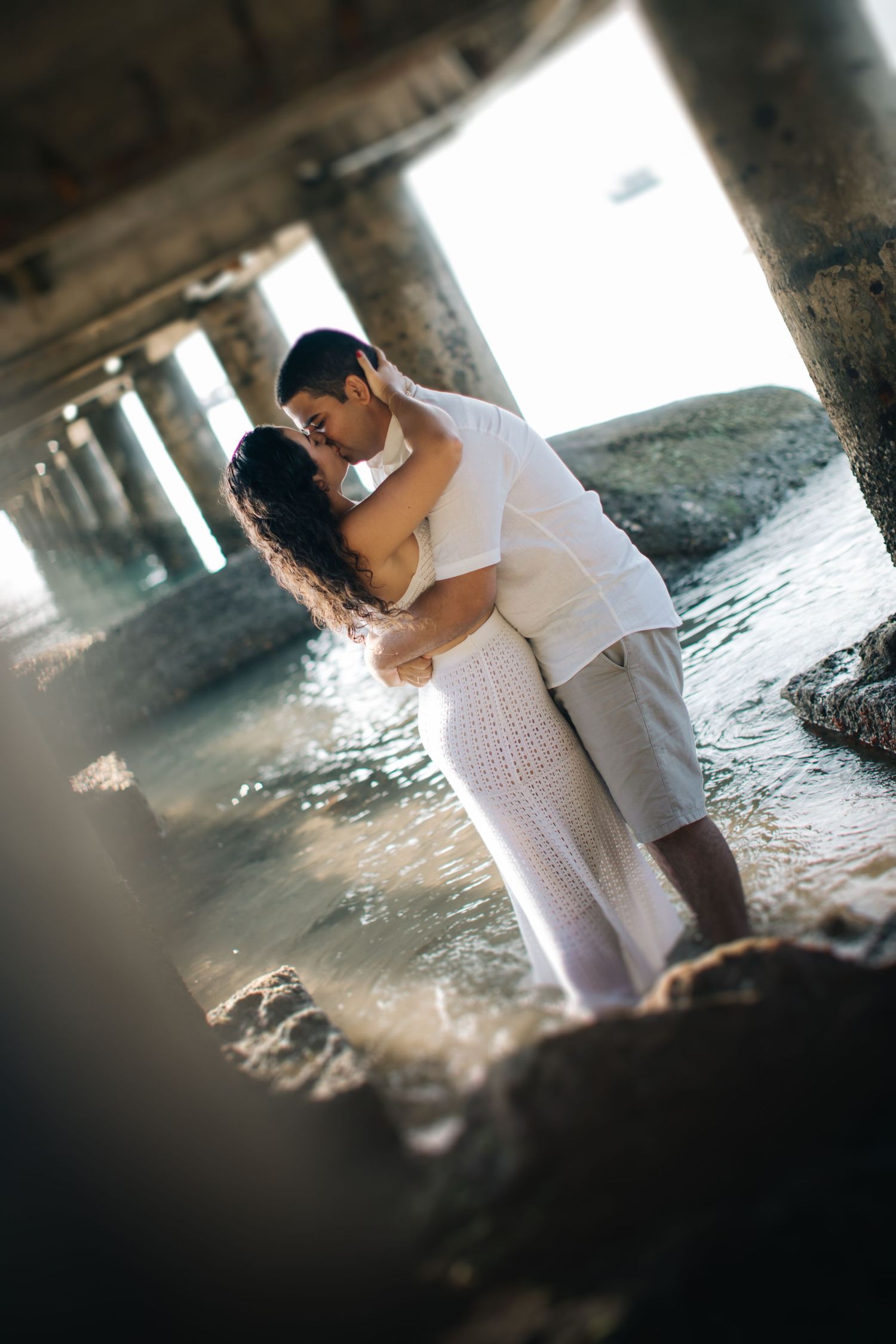 Romantic moment captured under pier with couple in white outfits creating dramatic silhouette.
