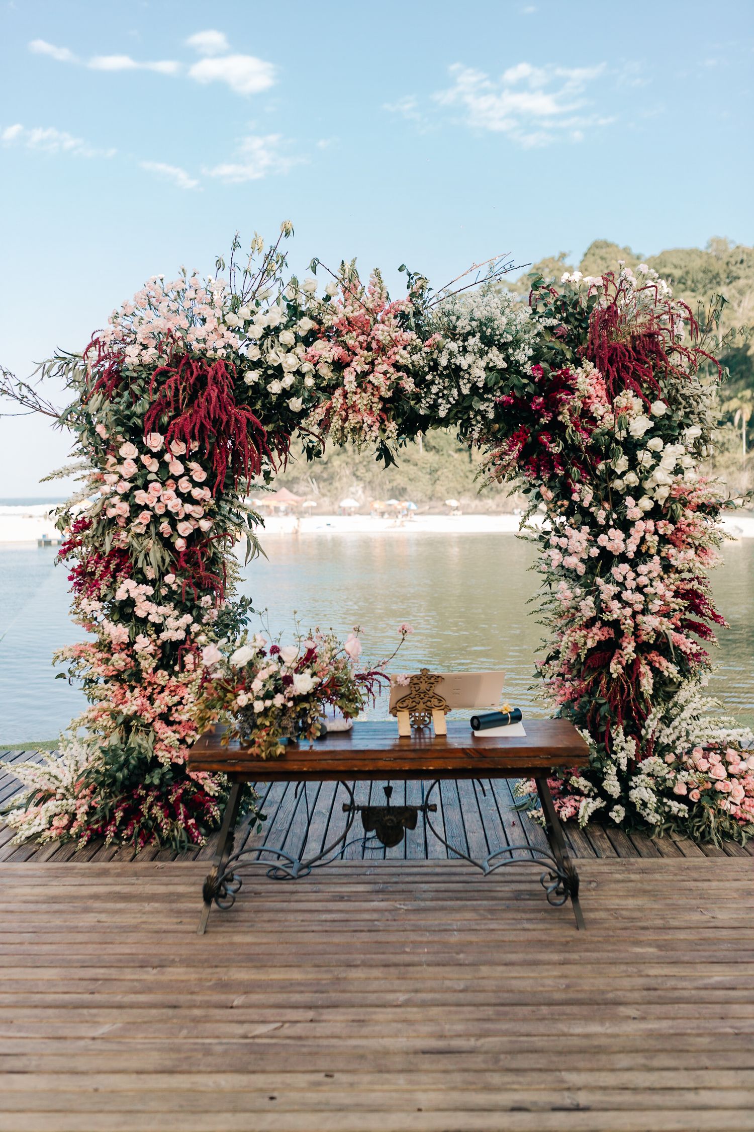 Floral arch with pink and burgundy flowers frames waterfront view on wooden deck.