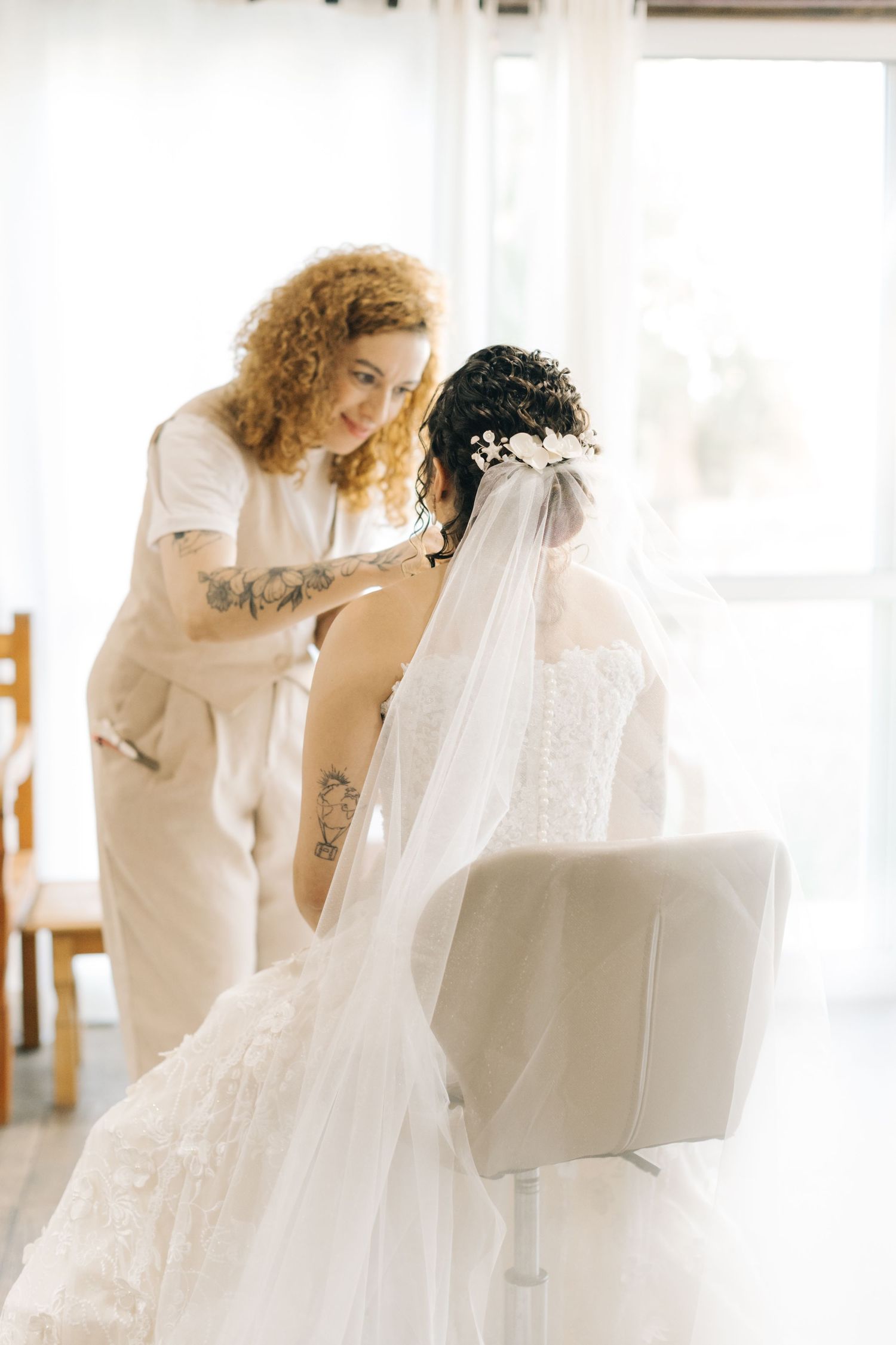 Sunlit wedding preparation showing stylist adjusting bride's hair and veil.