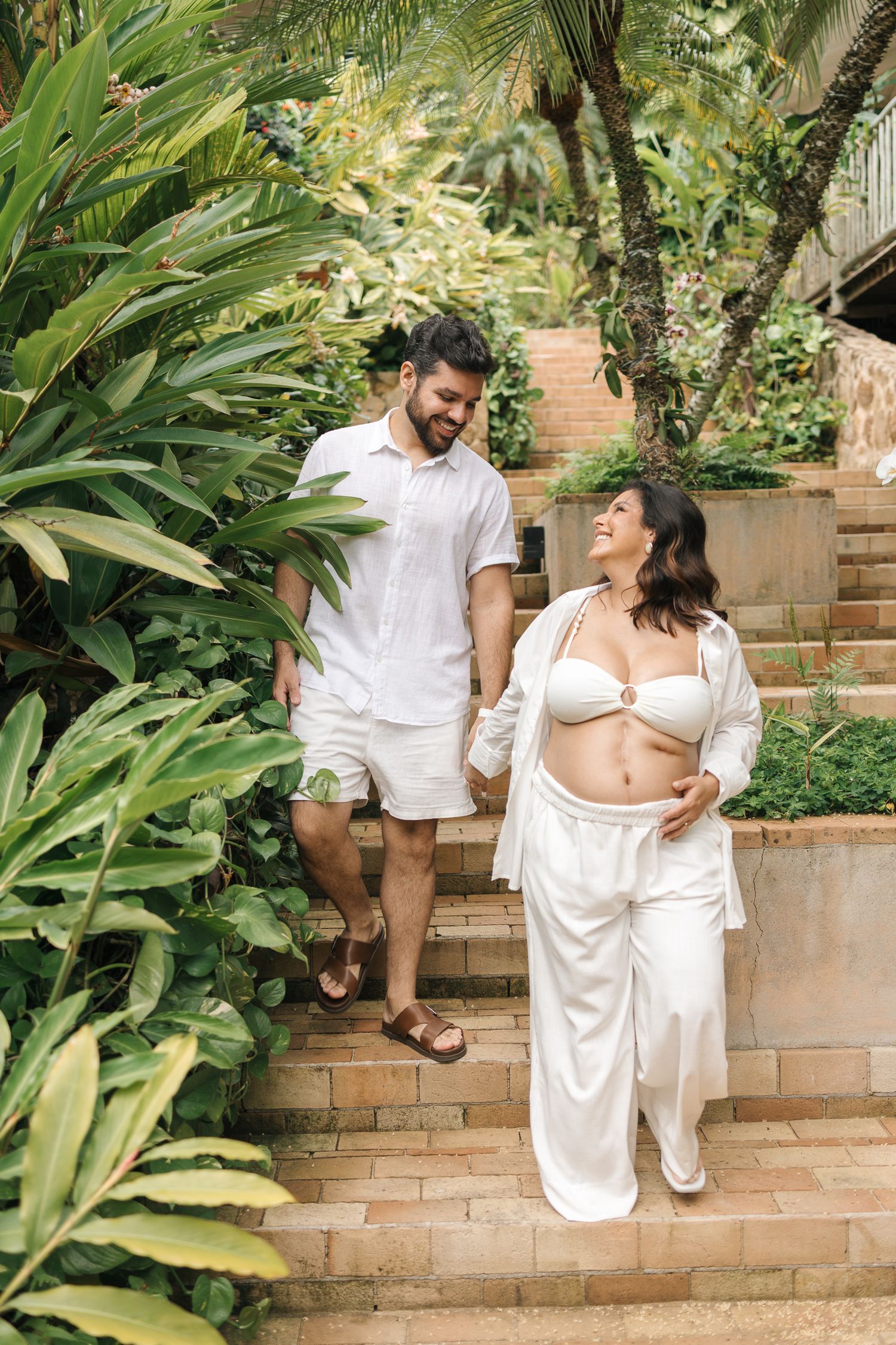 A couple in white outfits share romantic moments while sitting on outdoor brick steps surrounded by tropical plants.