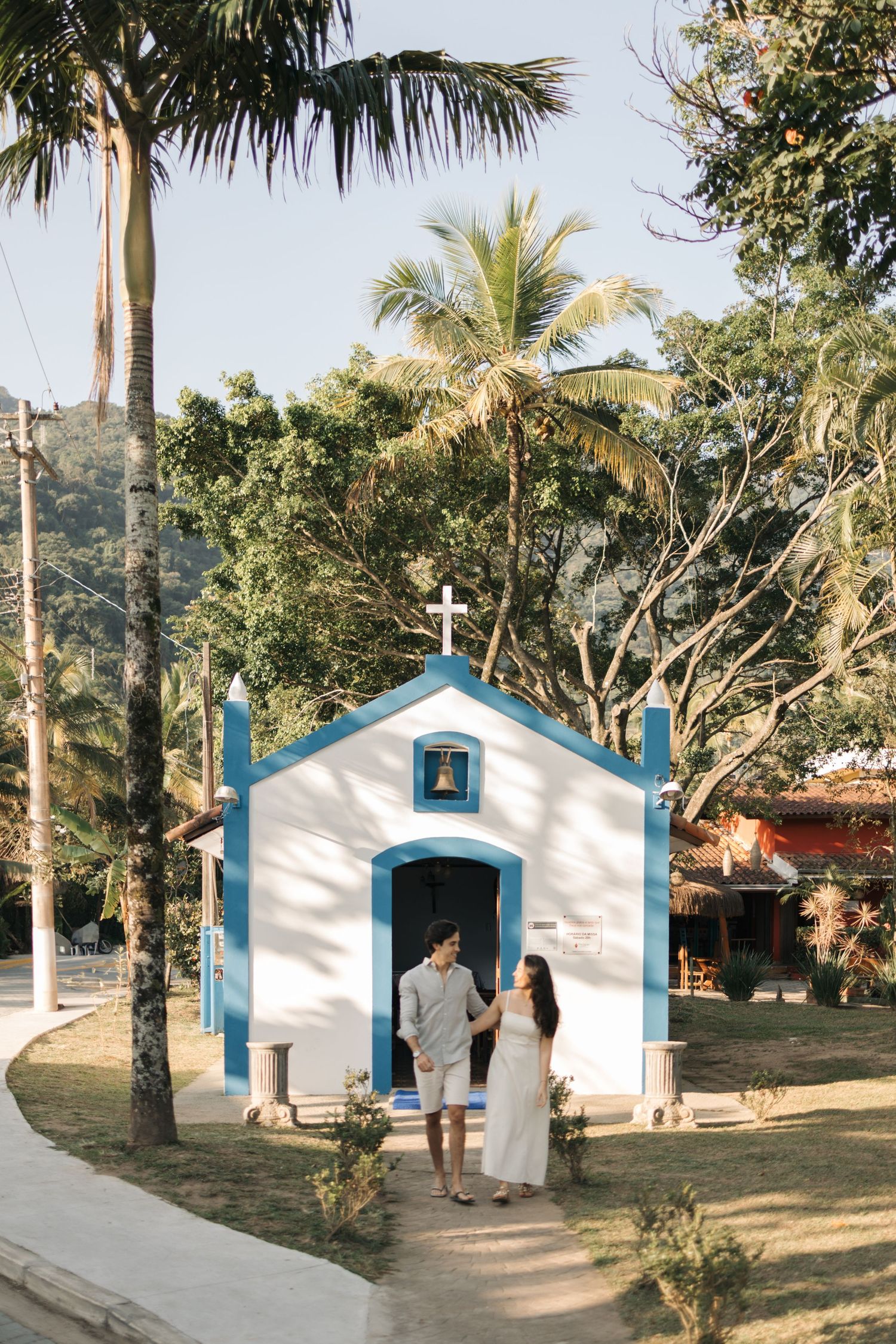 Small white chapels with blue trim line a palm-fringed path at a tropical beachside church location.