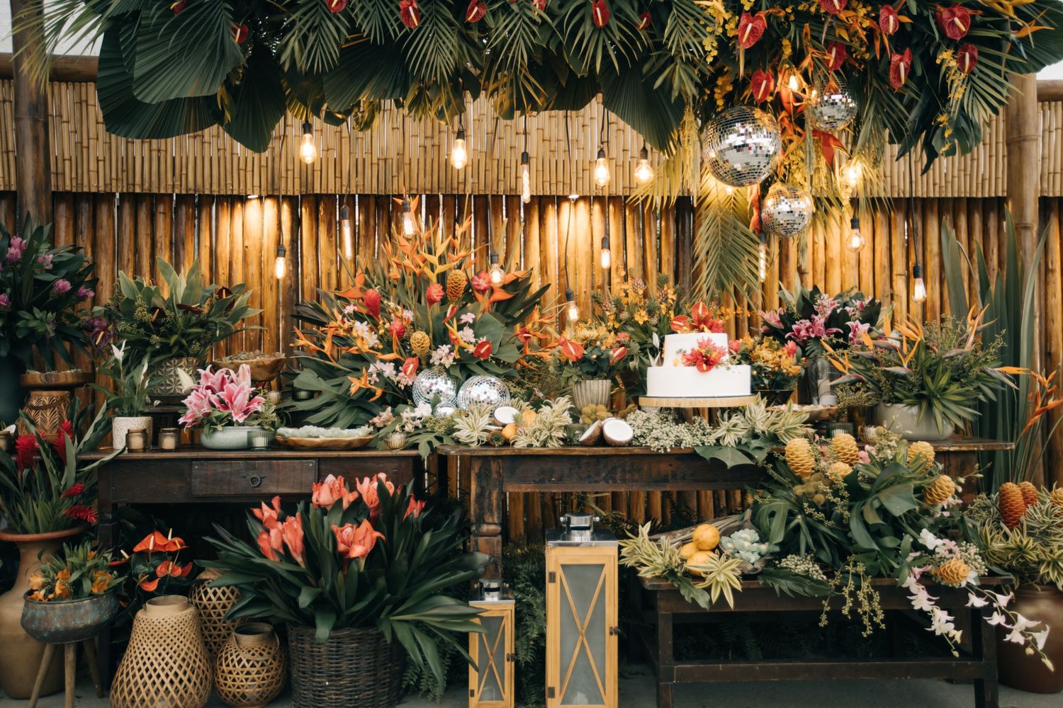 Elegant dessert display featuring tropical flowers, disco balls, and ambient lighting against bamboo wall.