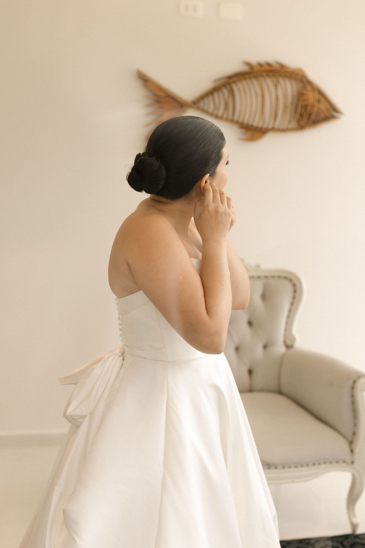 Person in white wedding gown adjusts jewelry while standing next to grey chair with fish artwork on wall.