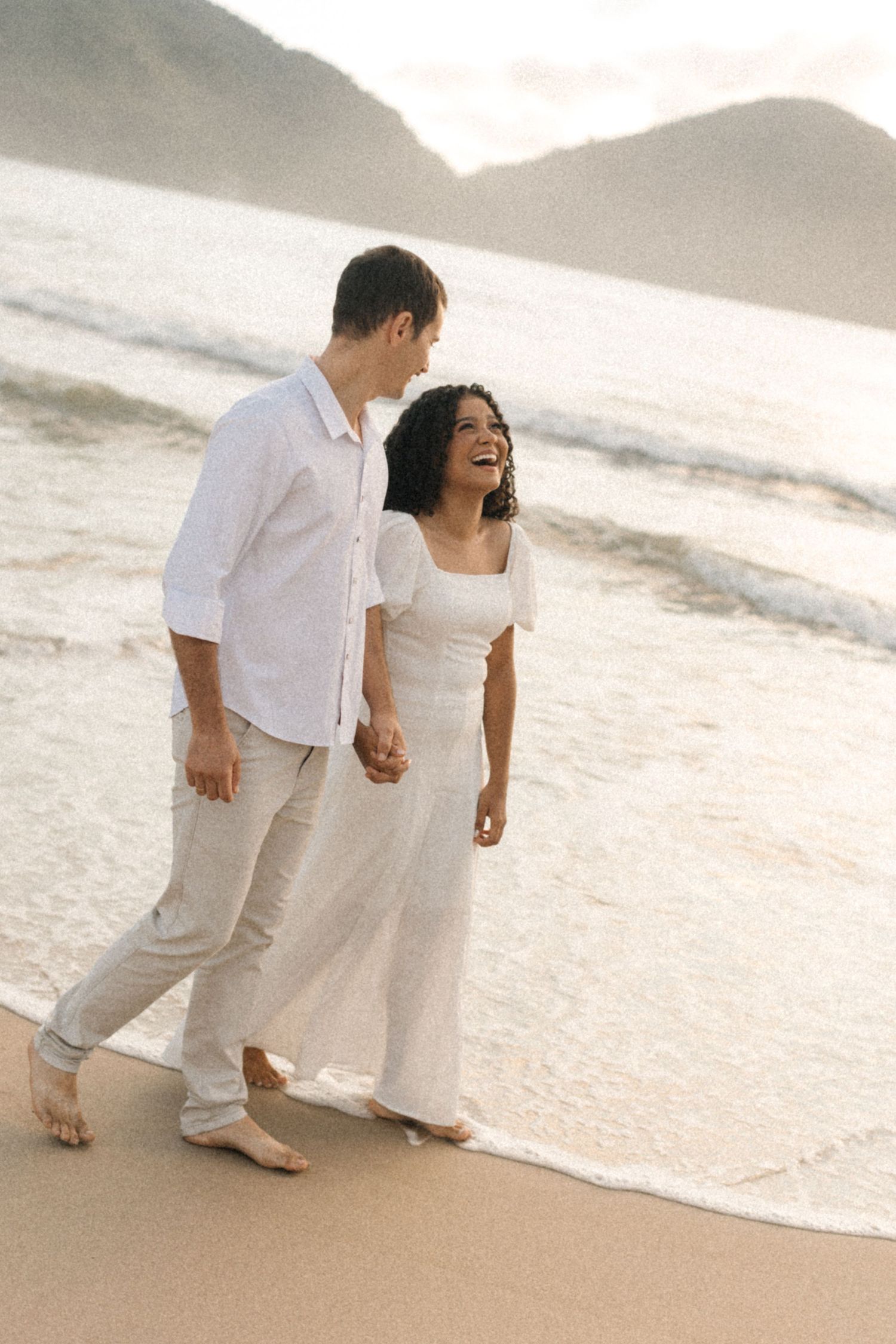 A couple in white attire walks together on a serene beach at sunset, creating romantic silhouettes in the sand.