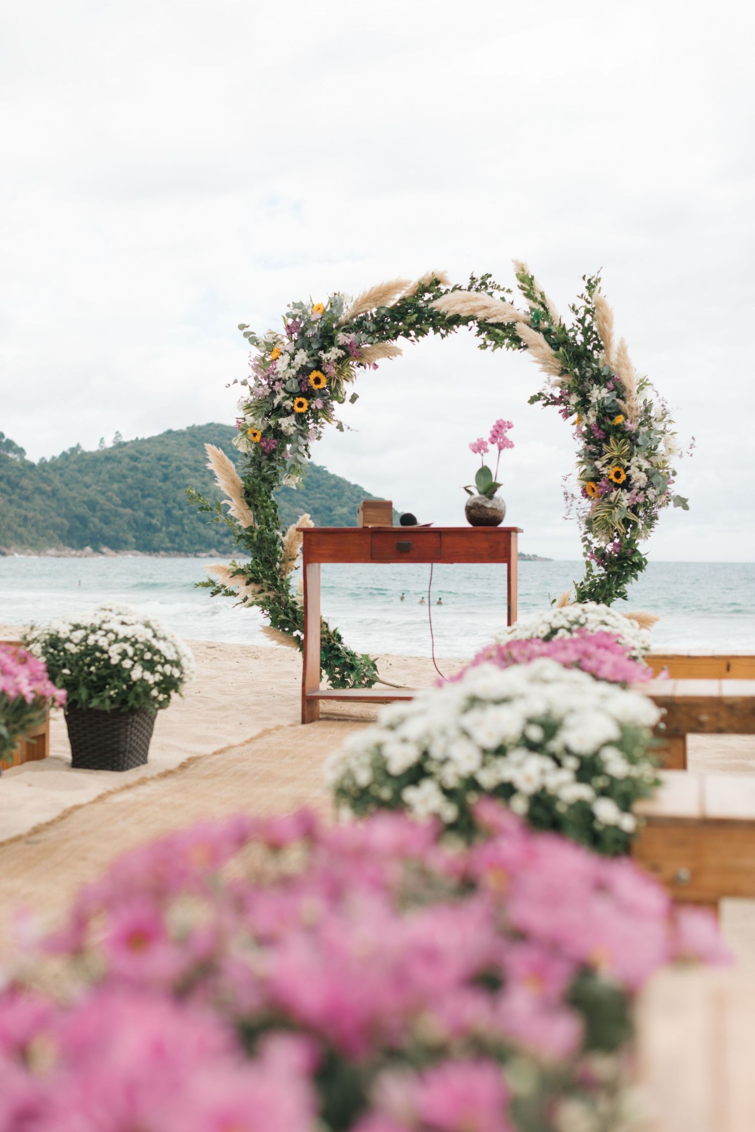 Beautiful floral wedding arch with pink and white flowers frames a wooden table on a serene beach setting.