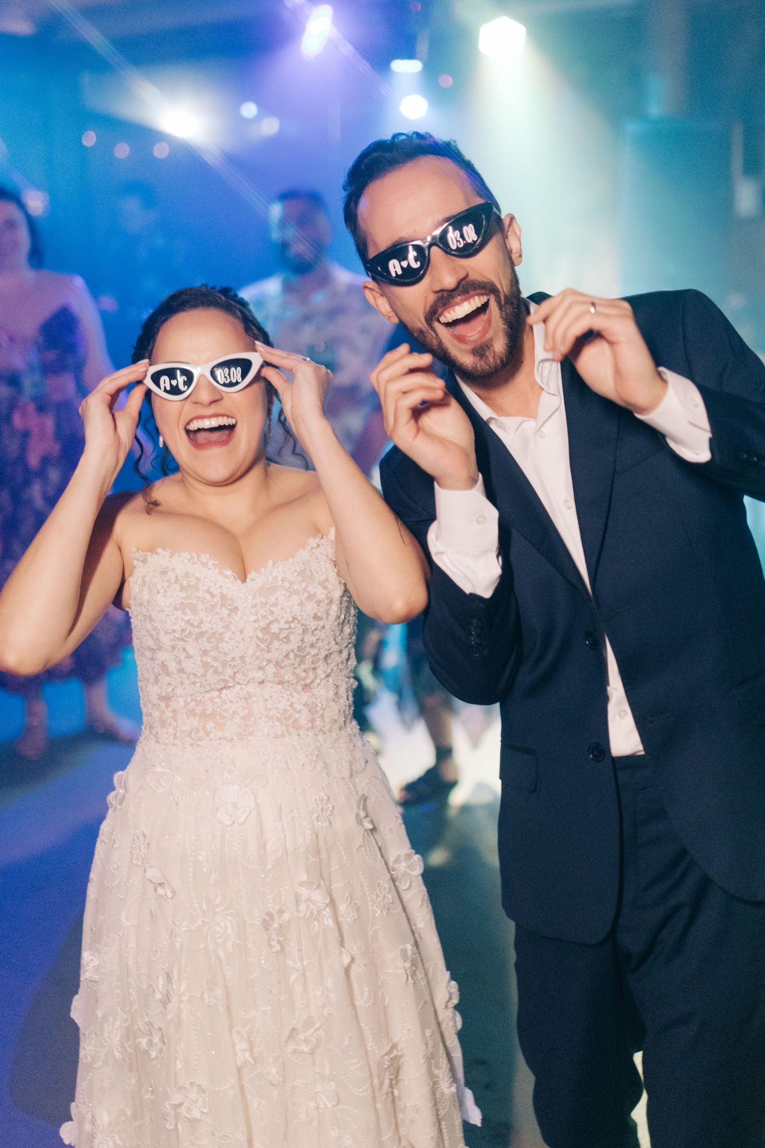 Happy couple dancing and posing with sunglasses at a reception with colorful lighting effects.