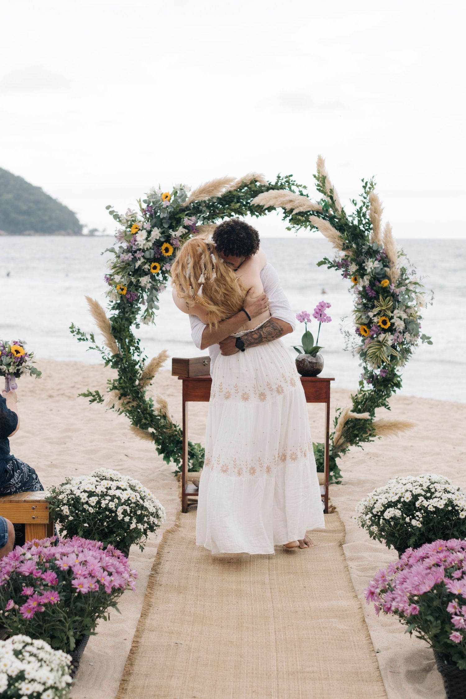 Newlyweds embrace under a decorative floral arch on a beach with pink and white flowers lining the aisle.