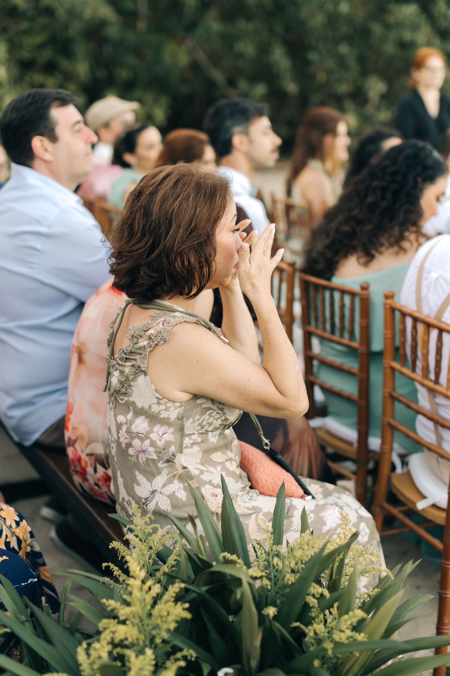 Wedding guest in a floral dress reacts emotionally during an outdoor ceremony among green foliage and wooden chairs.