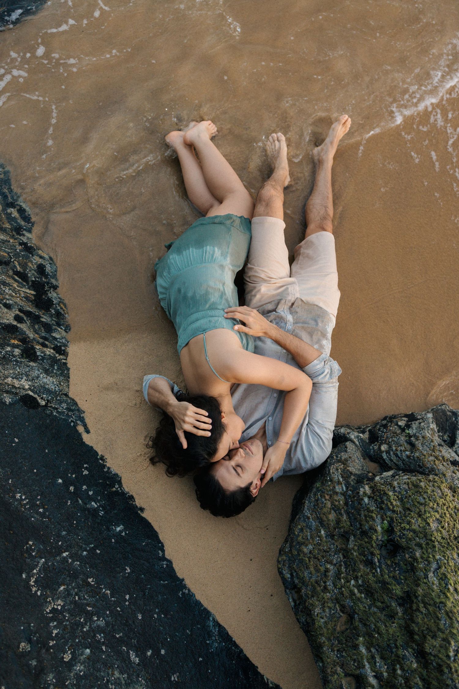 Couple lying together on wet sand next to ocean rocks during sunset.