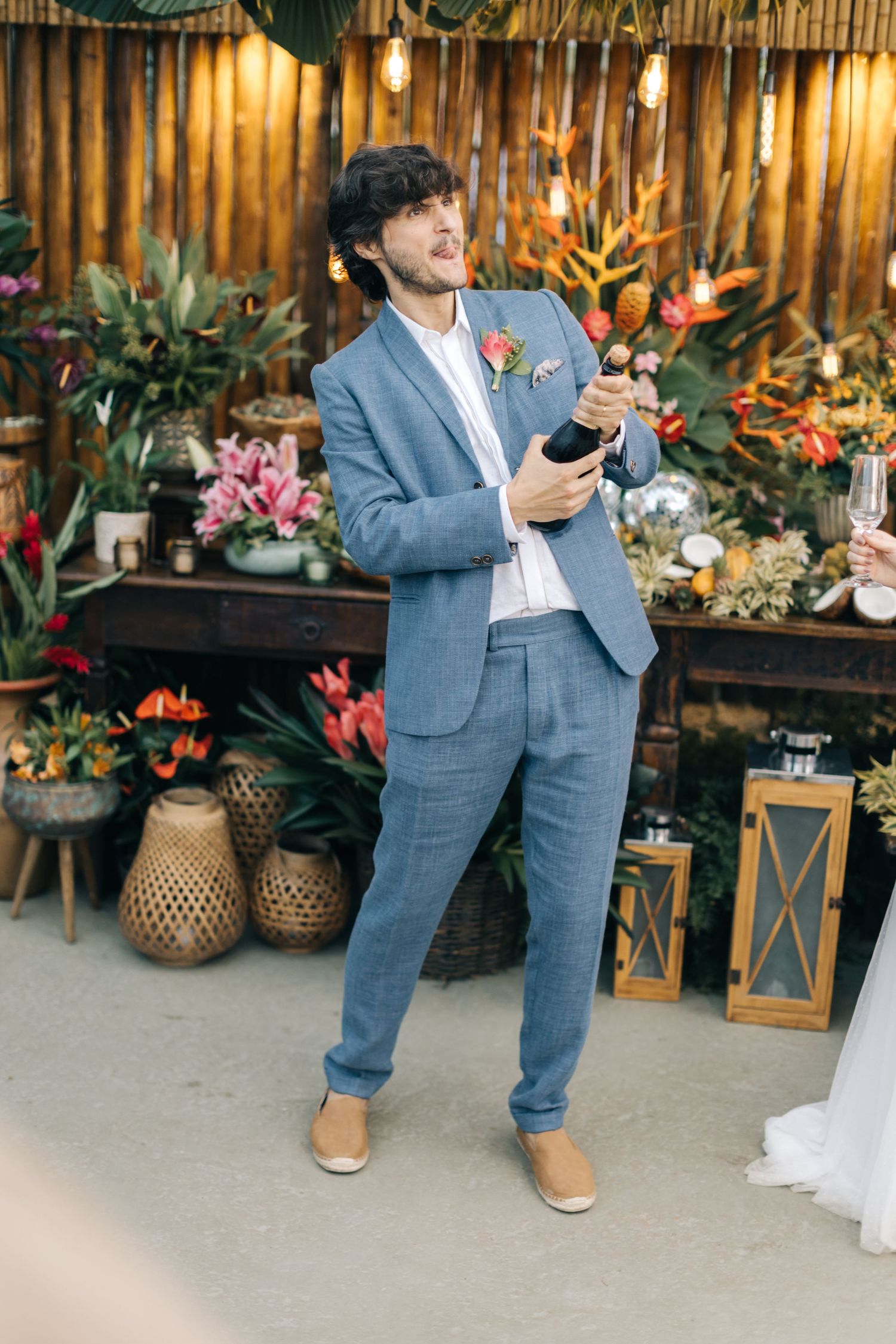 Groom in blue suit speaking at wedding reception with tropical decor and lanterns.