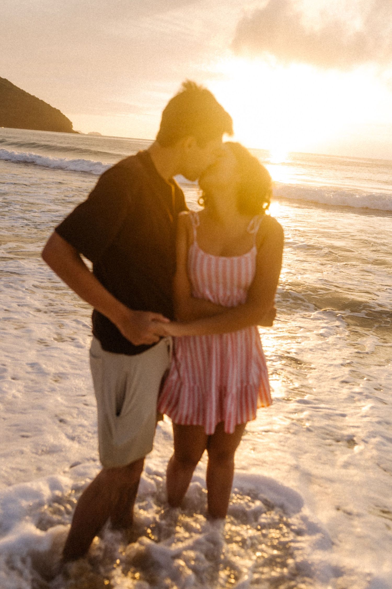 Casal se abraça em uma praia ao pôr do sol com ondas batendo em seus pés enquanto a luz dourada ilumina a cena romântica.