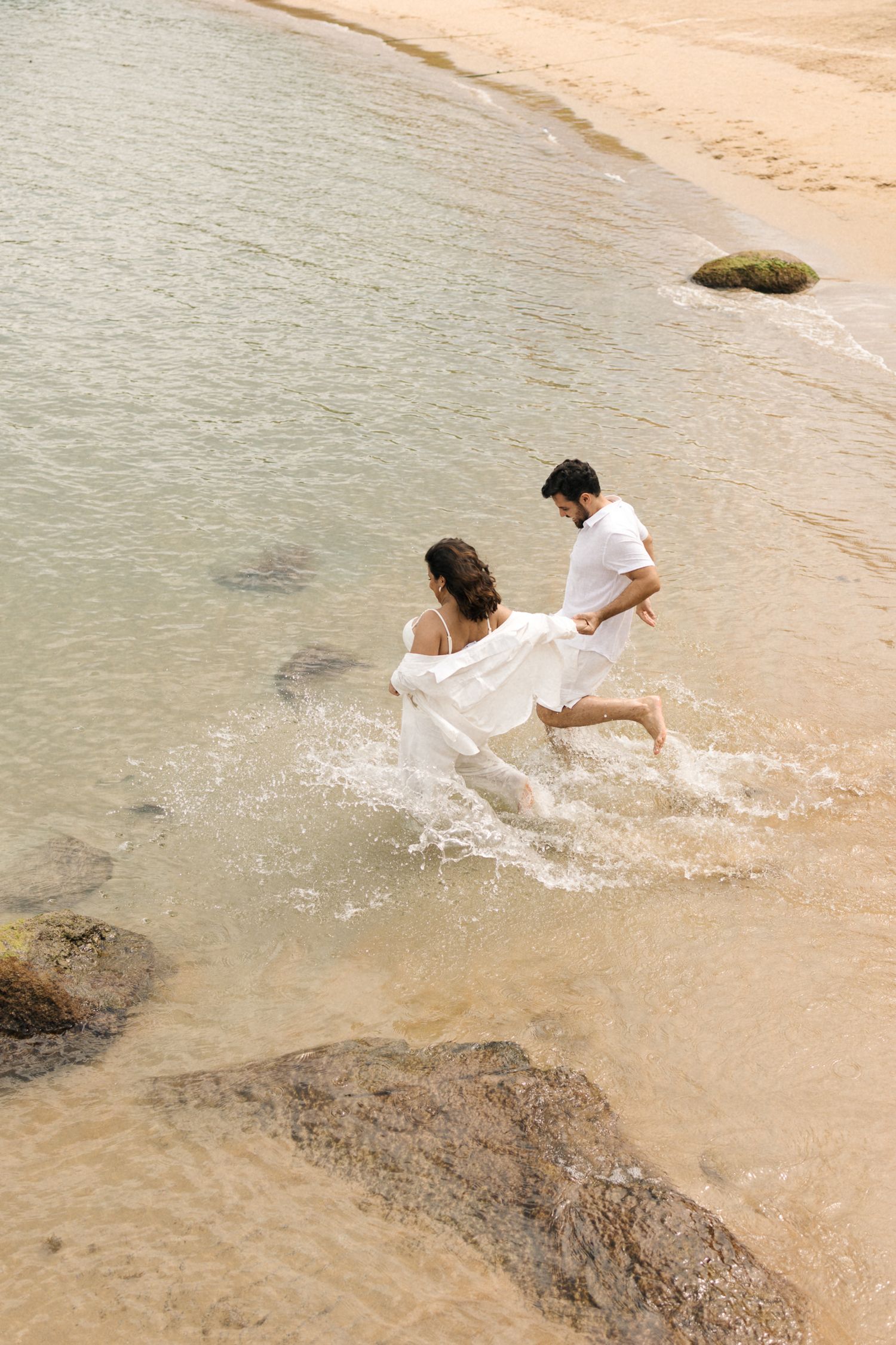 Two people play and splash in shallow coastal waters near rocky formations on a sandy beach.