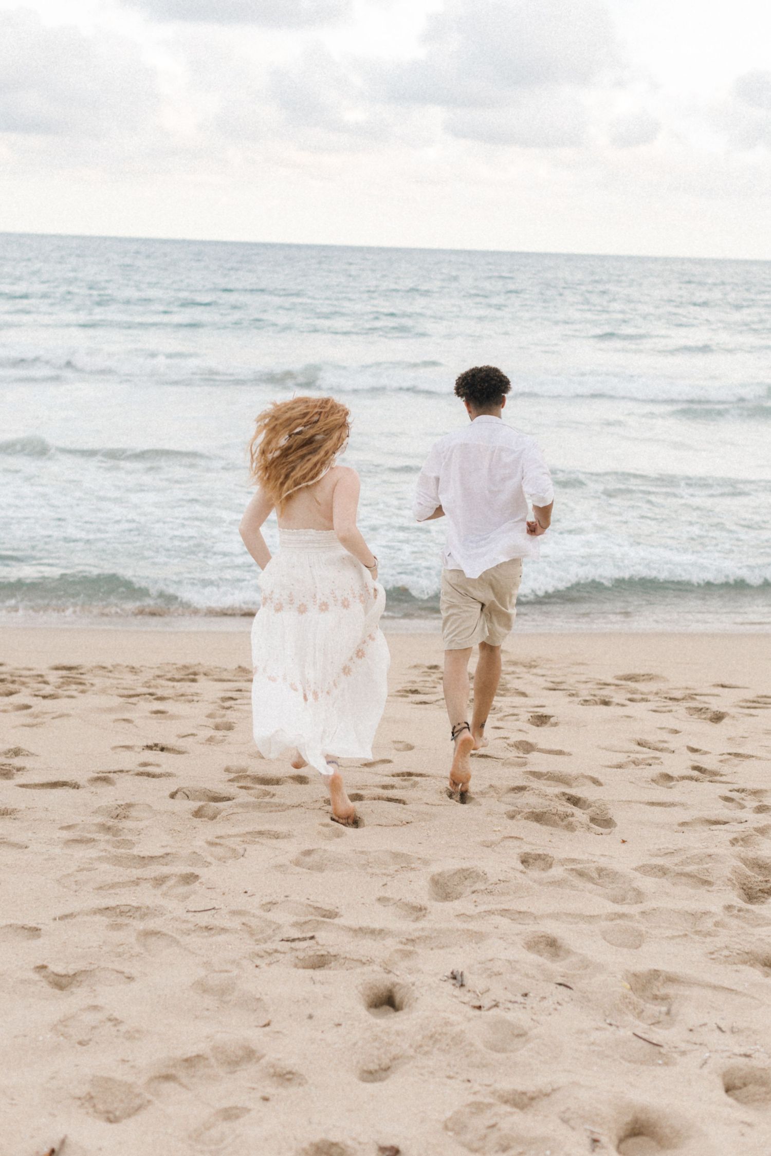 A joyful couple runs along the beach together in white summer clothing.