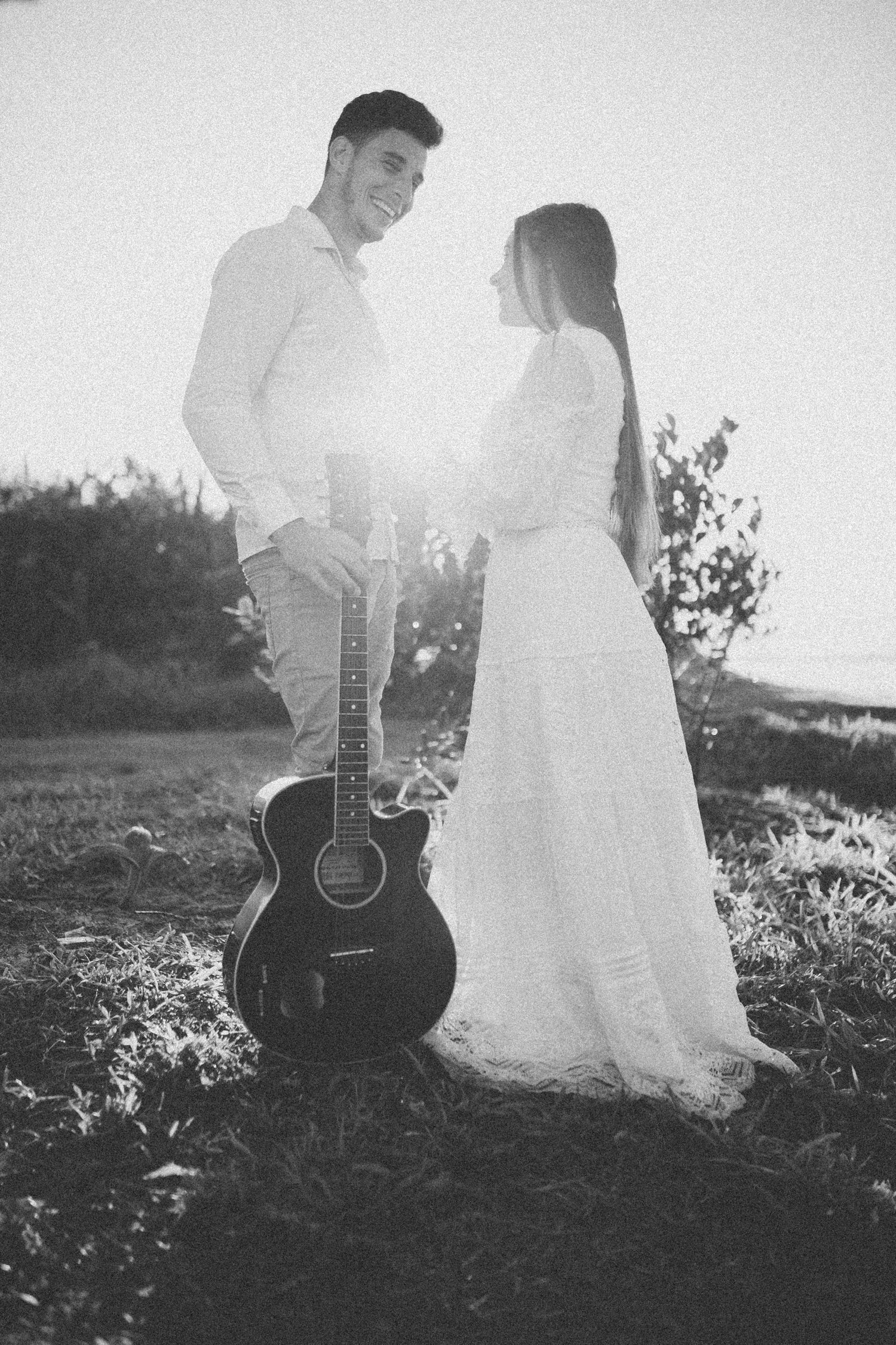 Black and white portrait of couple with guitar standing against sunset backdrop.