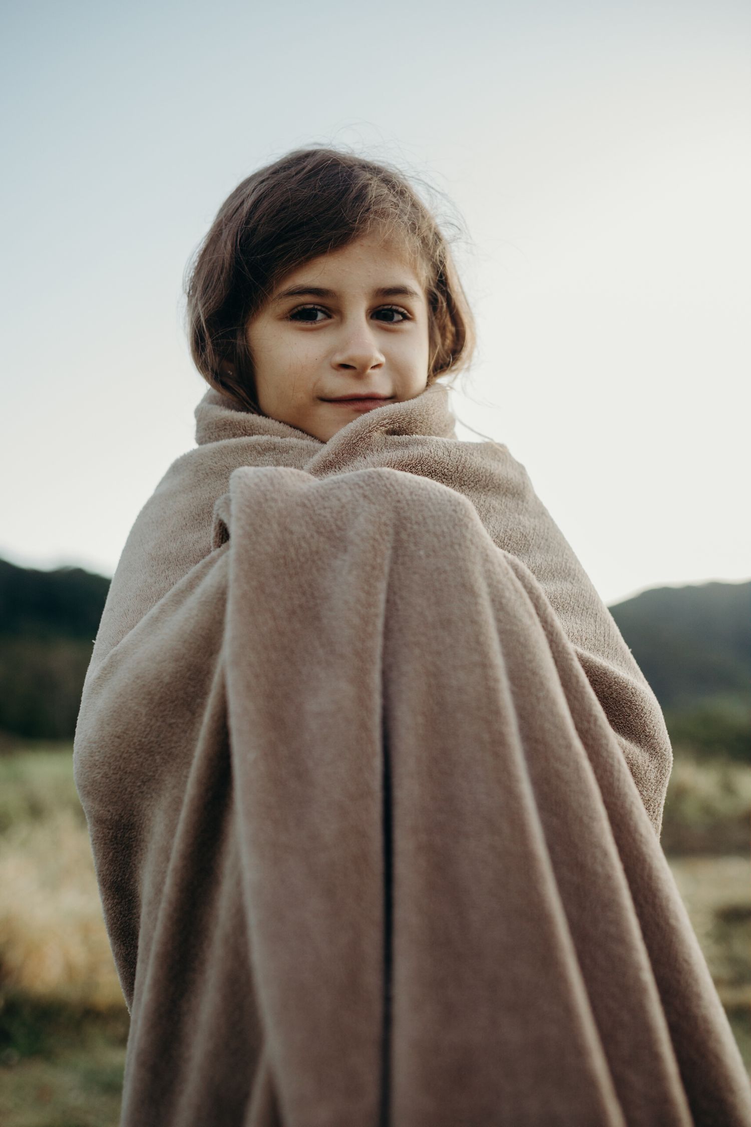 A child wrapped in a cozy beige blanket stands outdoors against a blurred background.