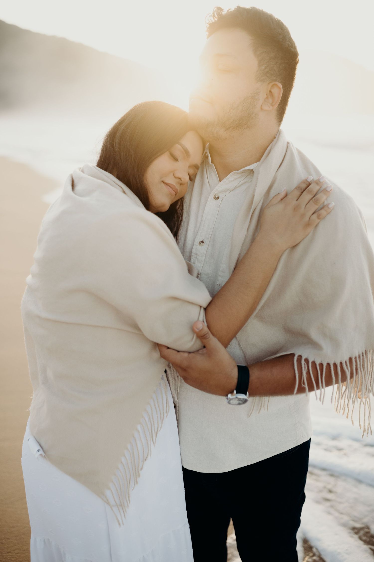 Couple embraces tenderly while wrapped in white shawl during beach sunset session.
