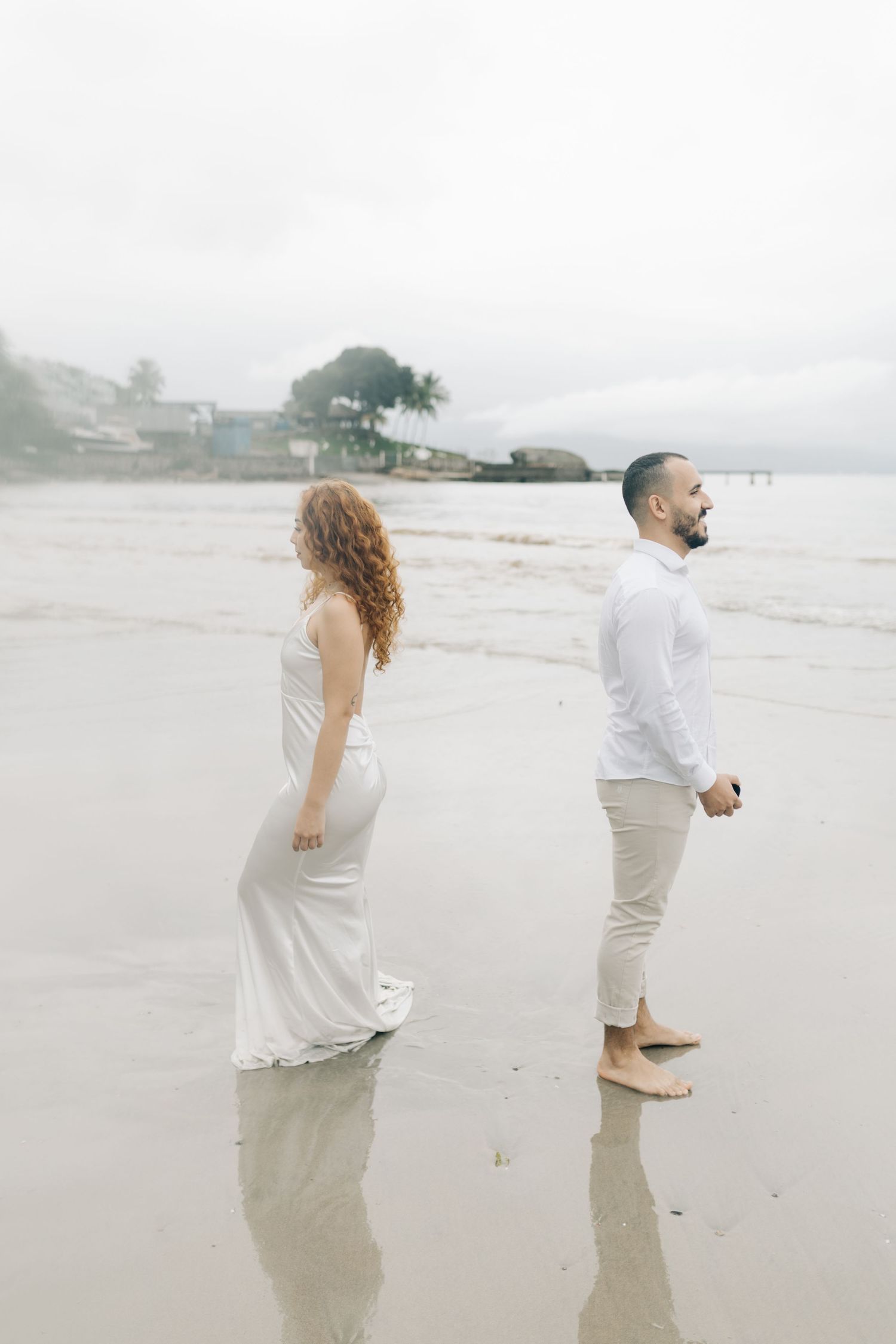Couple standing apart on reflective wet sand during foggy beach photoshoot.