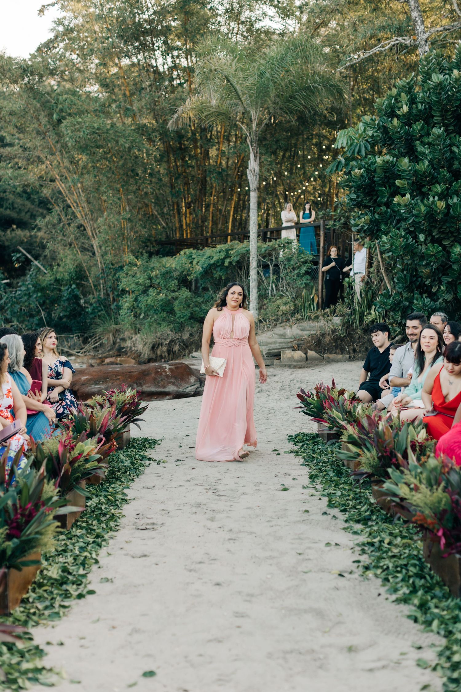 Person in pink dress walks down sandy aisle lined with tropical plants at outdoor ceremony.