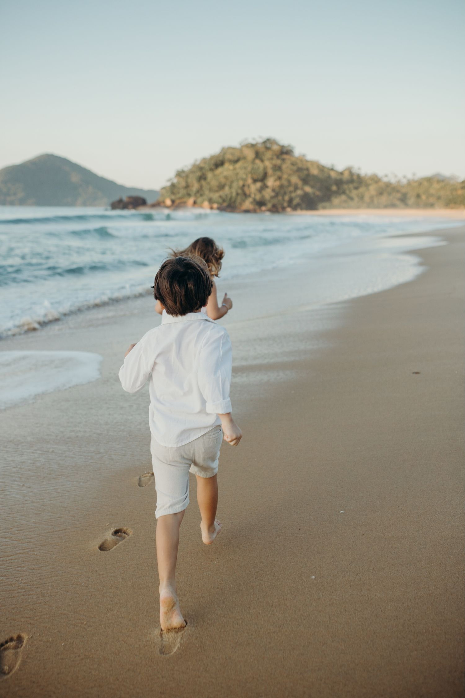 People in white clothing leave footprints while running along an empty beach with mountains in view.