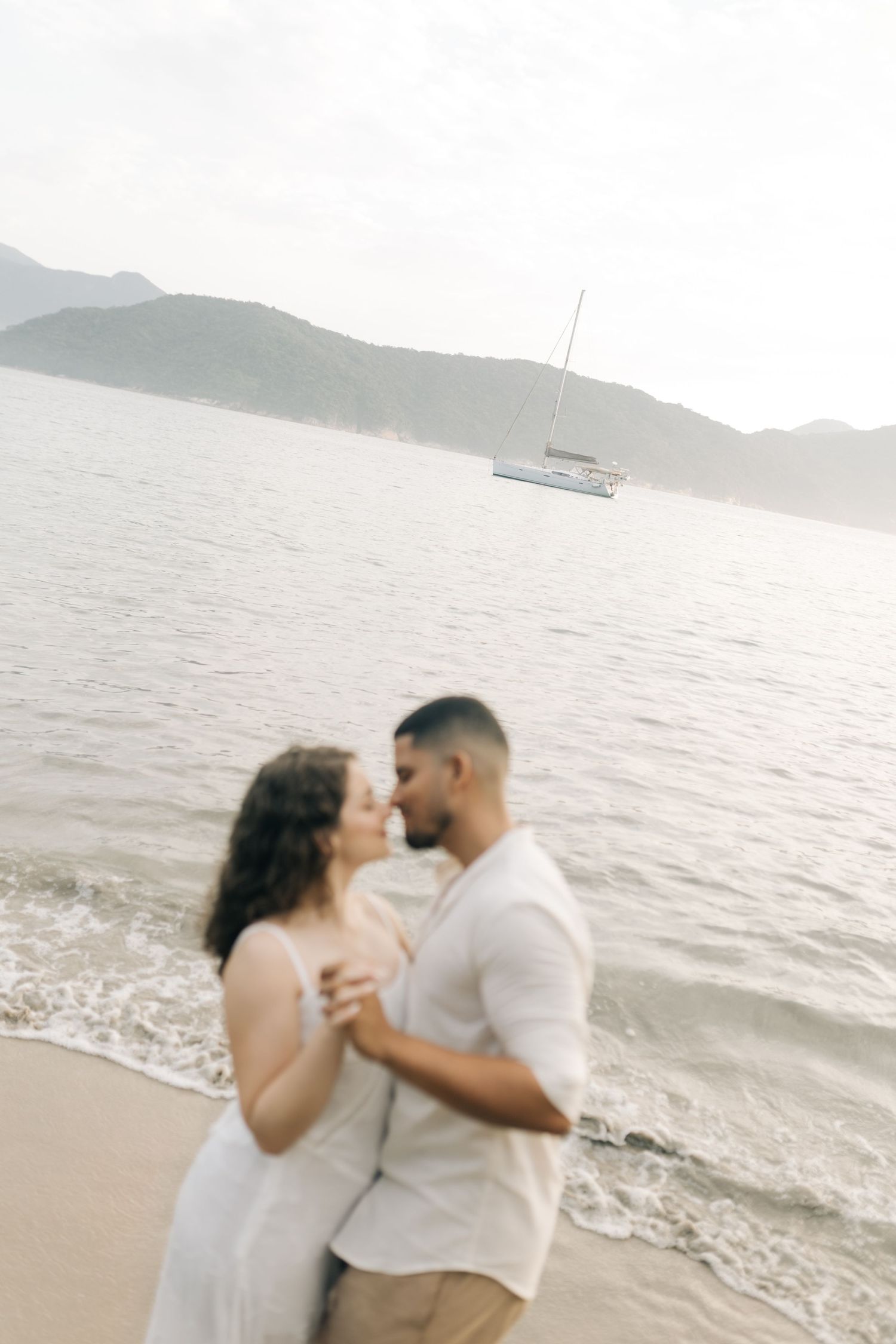 A couple shares an intimate moment on the beach with a sailboat visible in the distance.
