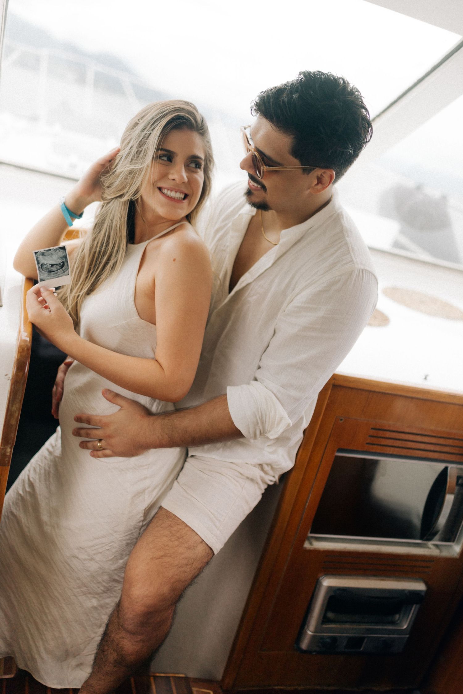 Couple in white outfits sharing an intimate moment on a boat, holding a photo.