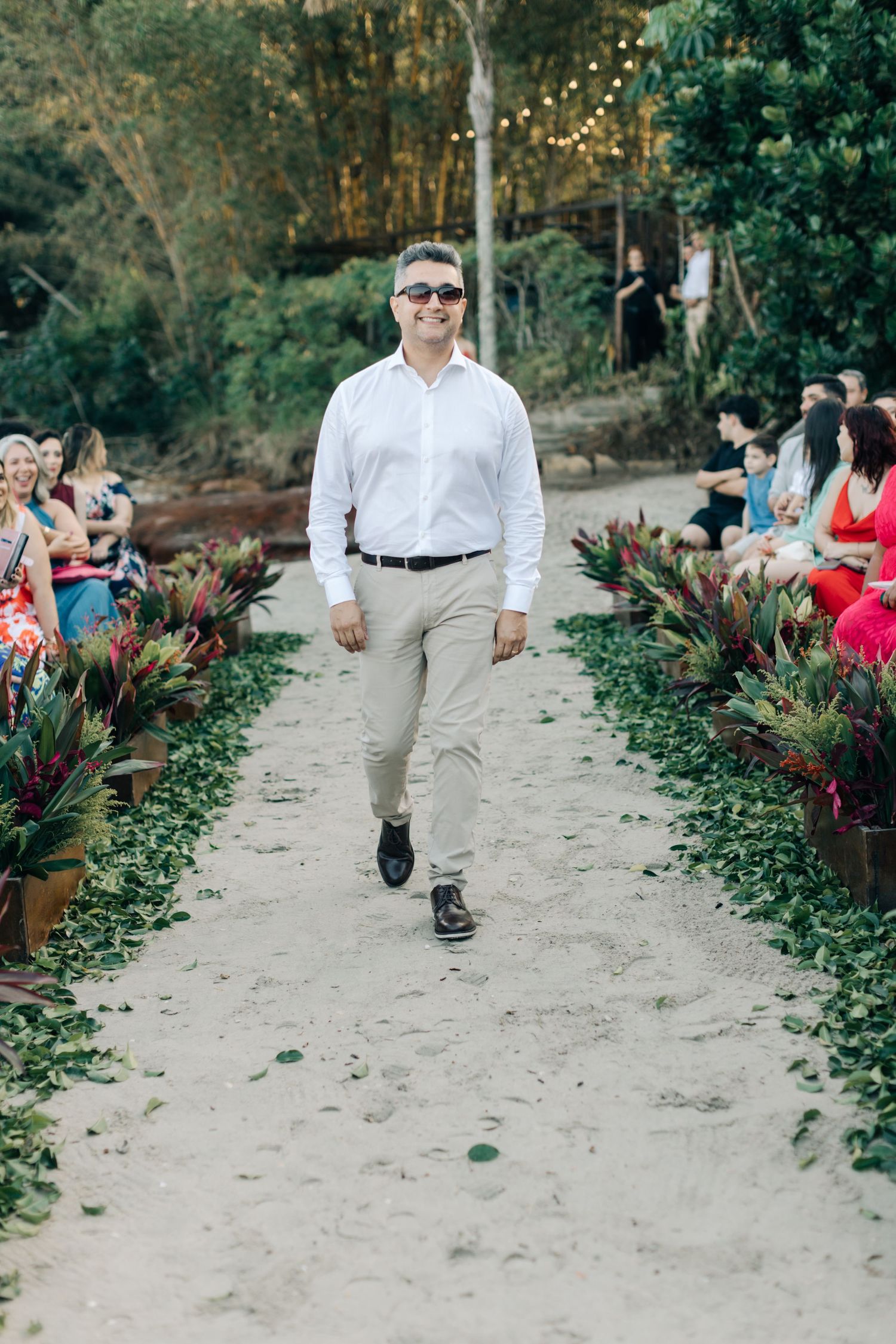 Wedding attendant in white dress shirt walks down outdoor ceremony aisle lined with tropical plants.
