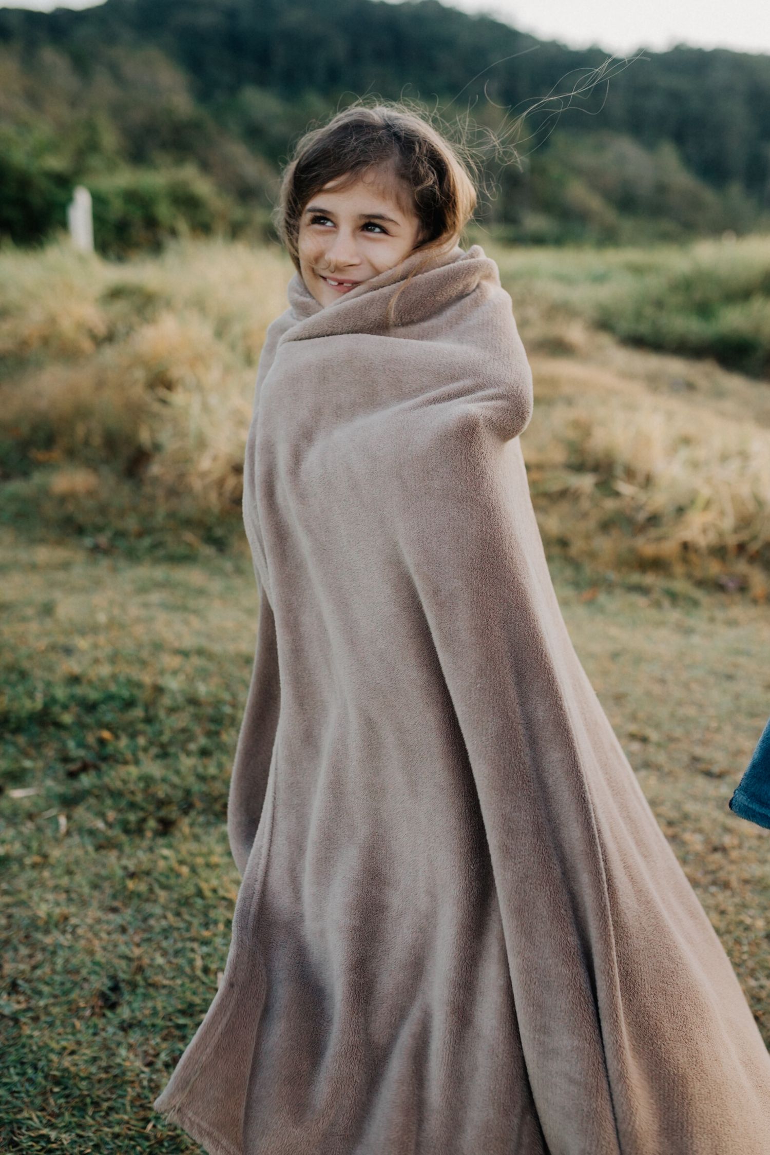 Person wrapped in a cozy beige blanket standing on a grassy hillside during golden hour.