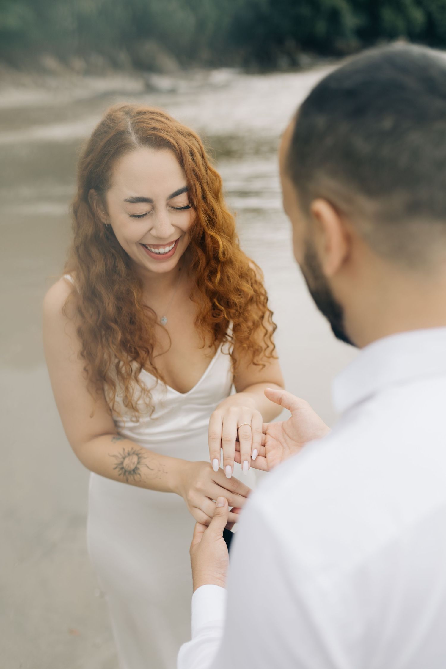Woman in white dress smiles joyfully while looking at engagement ring on beach.