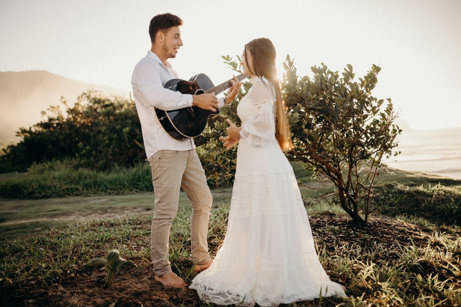Couple shares romantic moment with guitar on coastal hillside.
