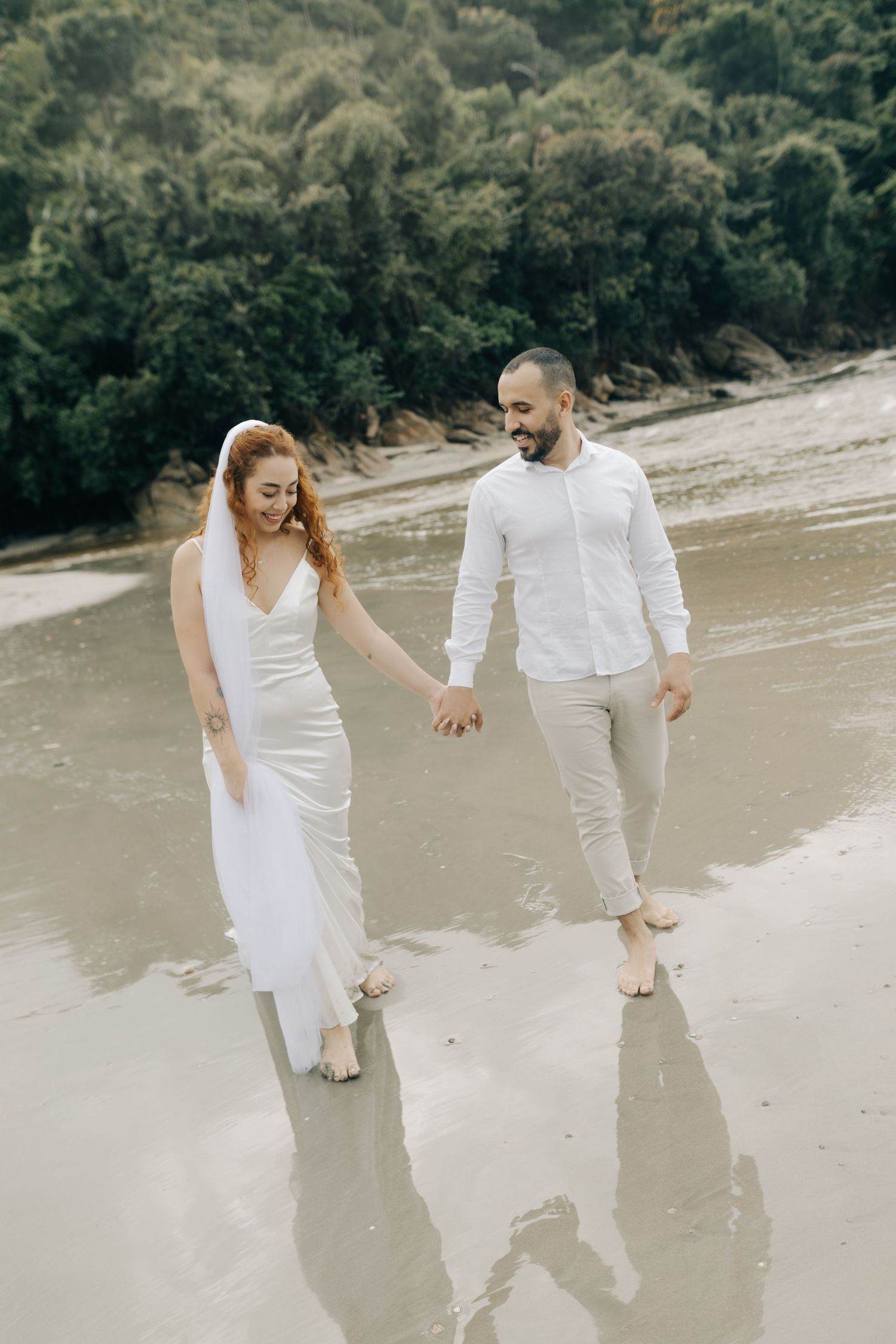 Couple in white outfits holding hands while walking on wet beach sand.