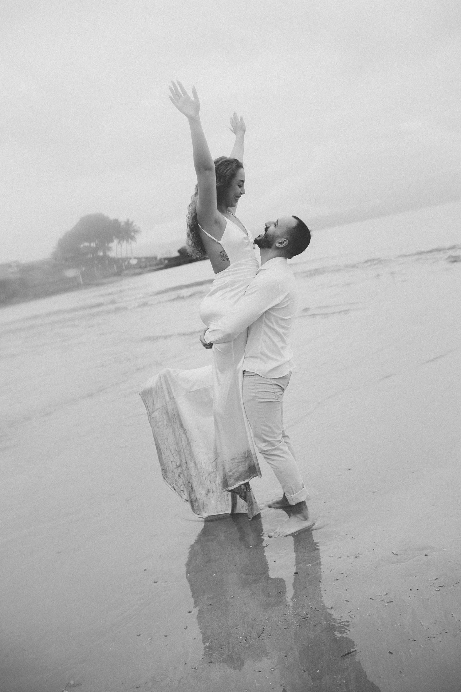 A joyful couple in white outfits dances and celebrates on a misty beach.
