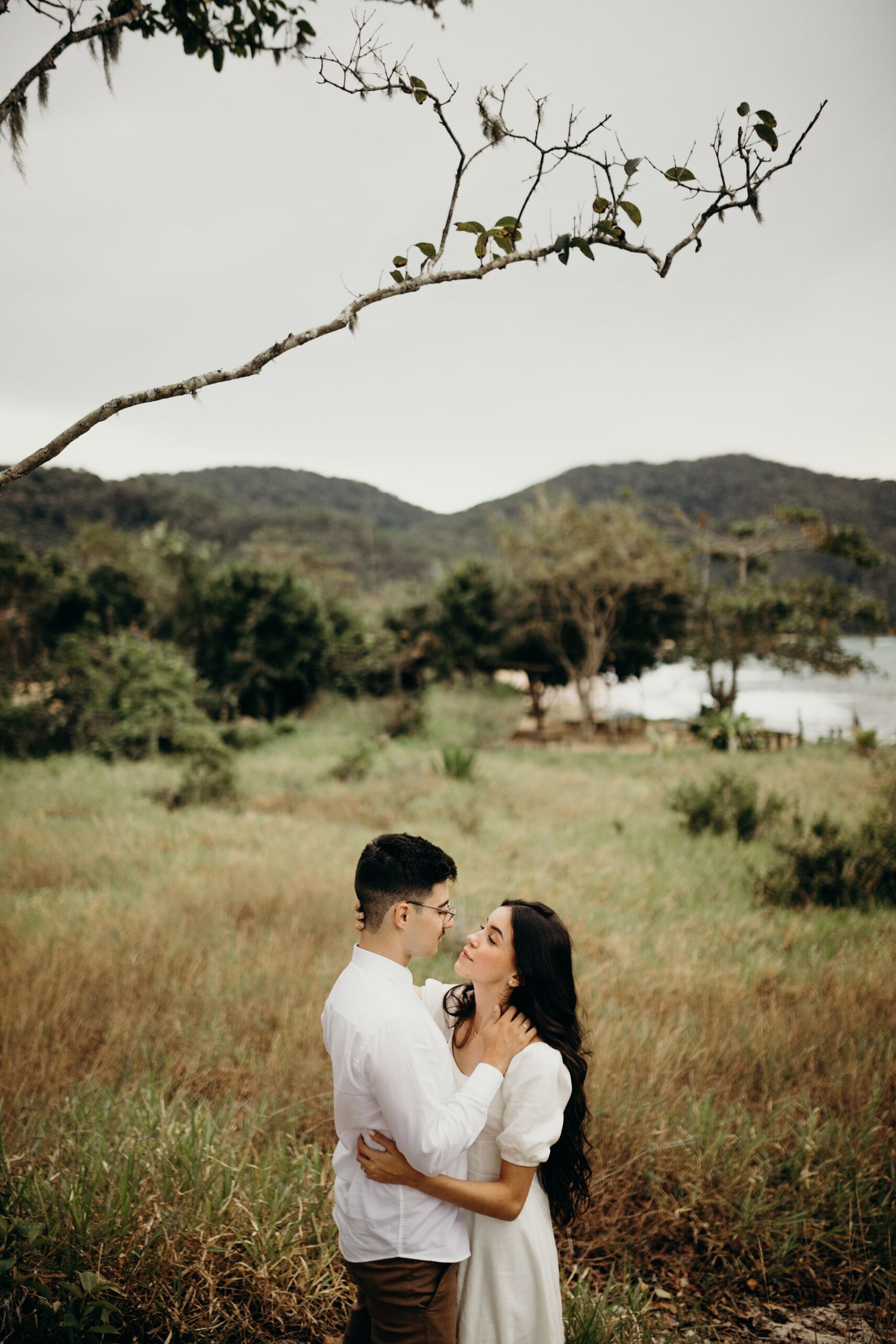 Couple shares an intimate moment in a grassy field with mountains and ocean in background.