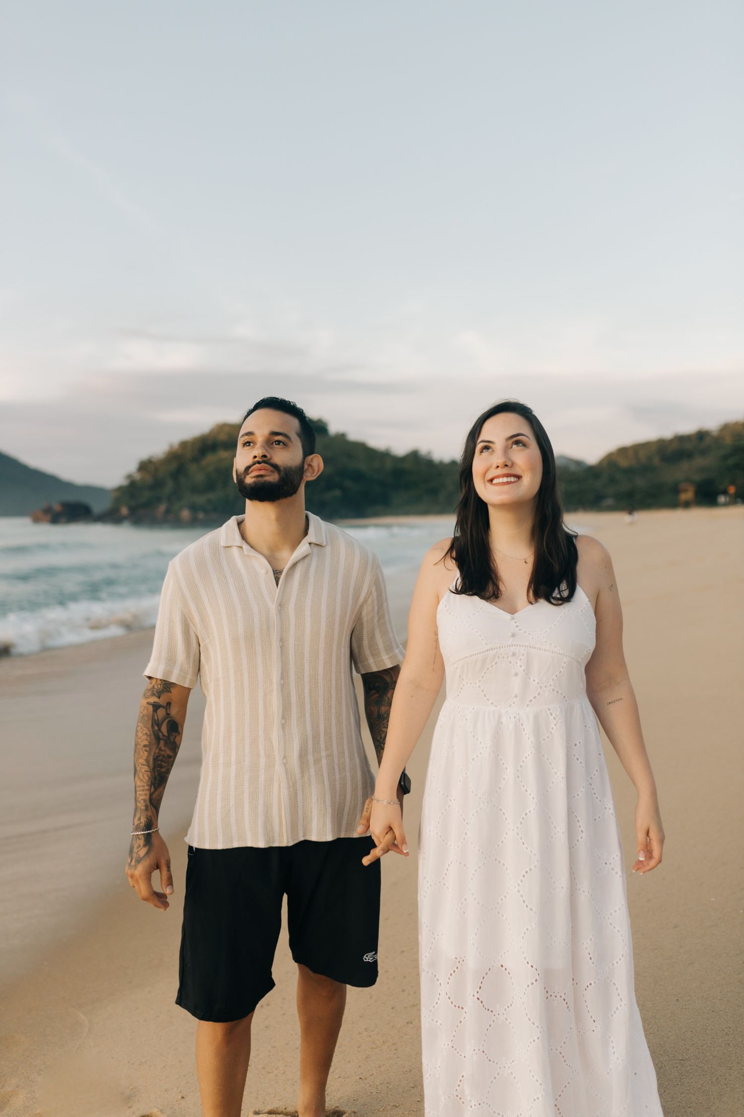 Couple in white and black outfits joyfully walks along sandy beach with ocean views.