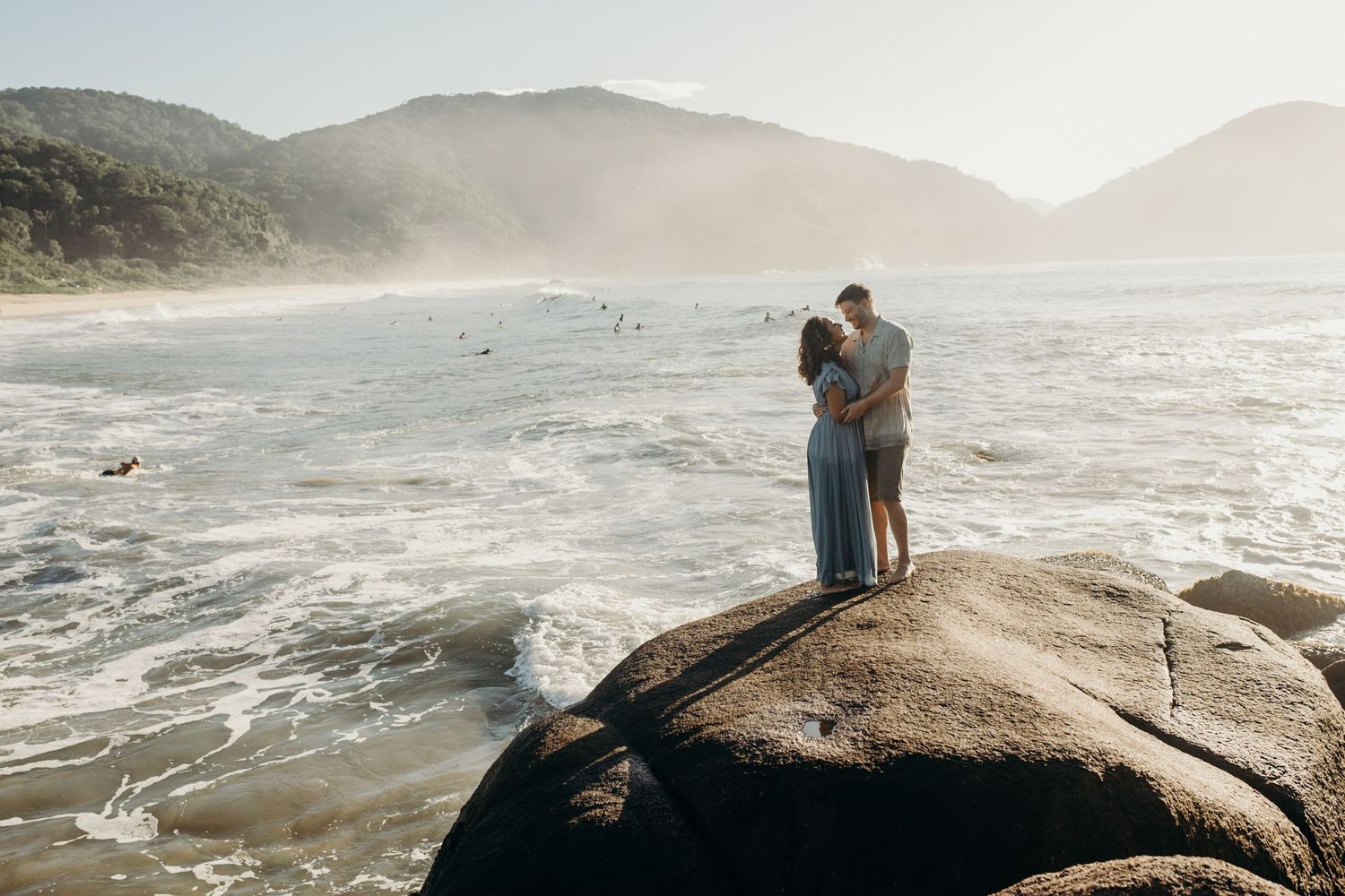 Couple standing on large boulder overlooking misty beach with mountains in distance.