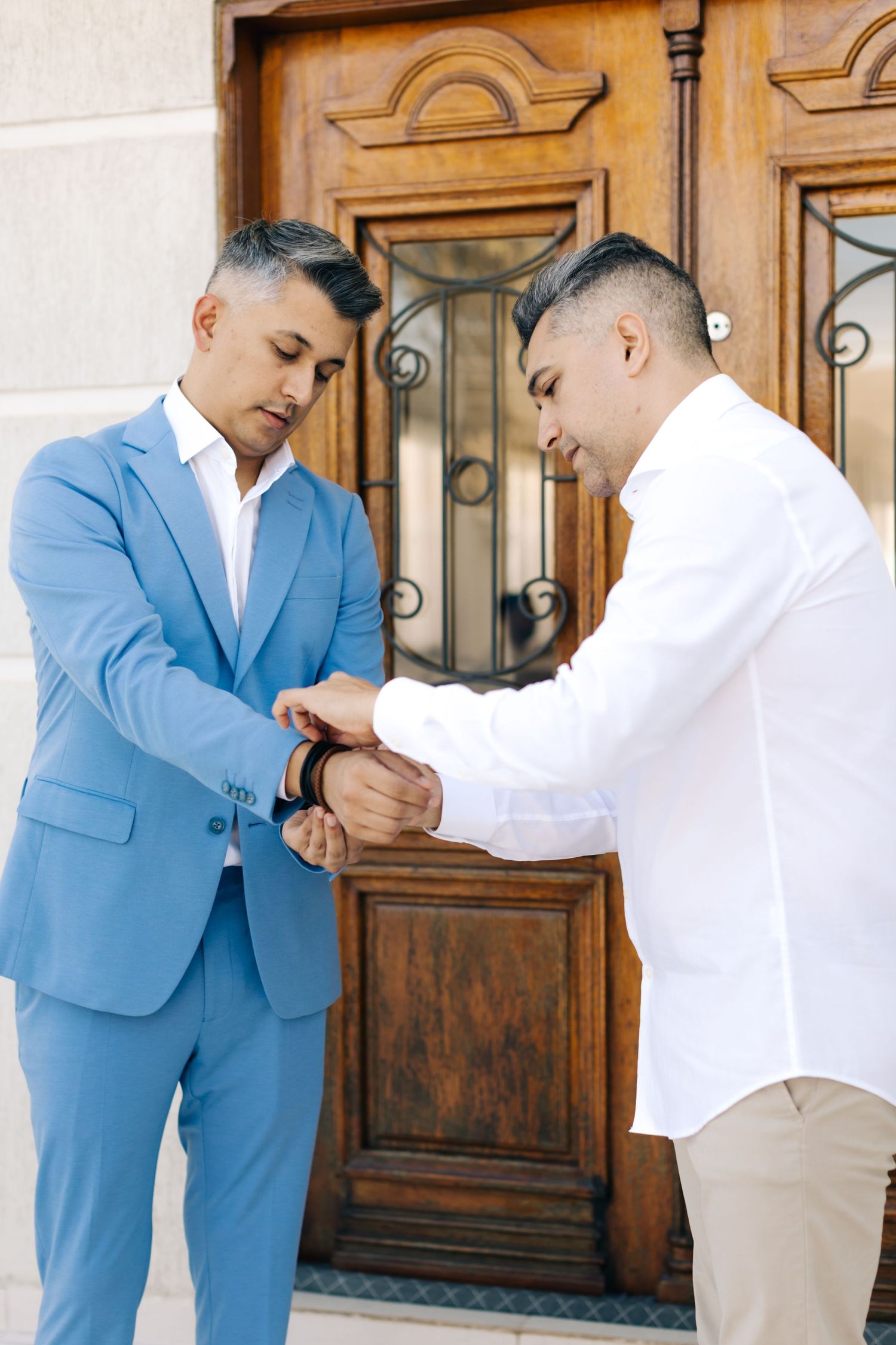 Two people in formal attire checking a watch or timepiece together in front of an ornate wooden door.
